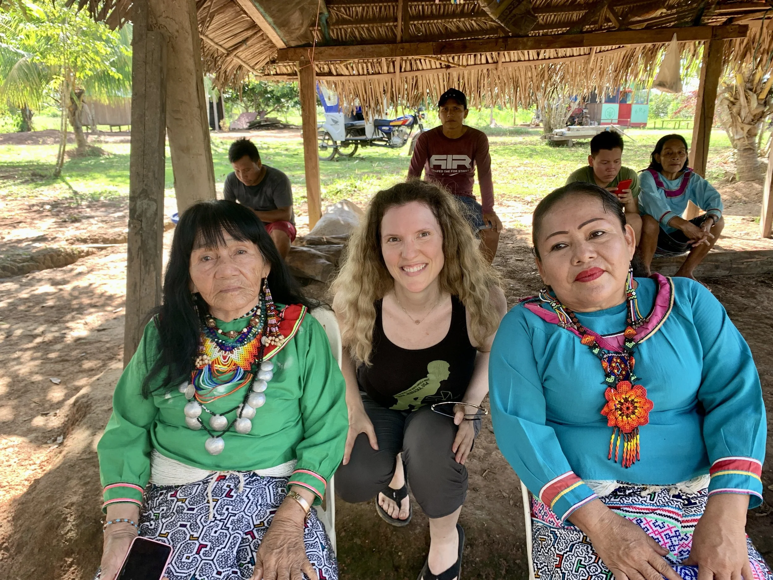 Group of five women and one man sitting outdoors under a thatched roof, some women wearing colorful traditional clothing and accessories, with a woman with curly hair smiling in the center, in a rural setting with trees and a motorcycle in the background.
