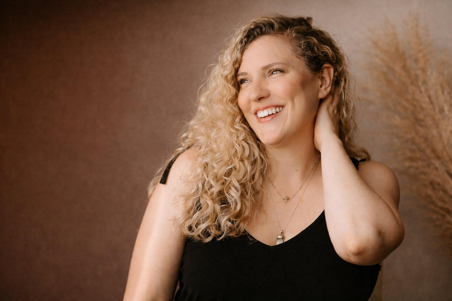 A woman with curly blonde hair smiling and touching her neck, wearing a black top and layered necklaces.