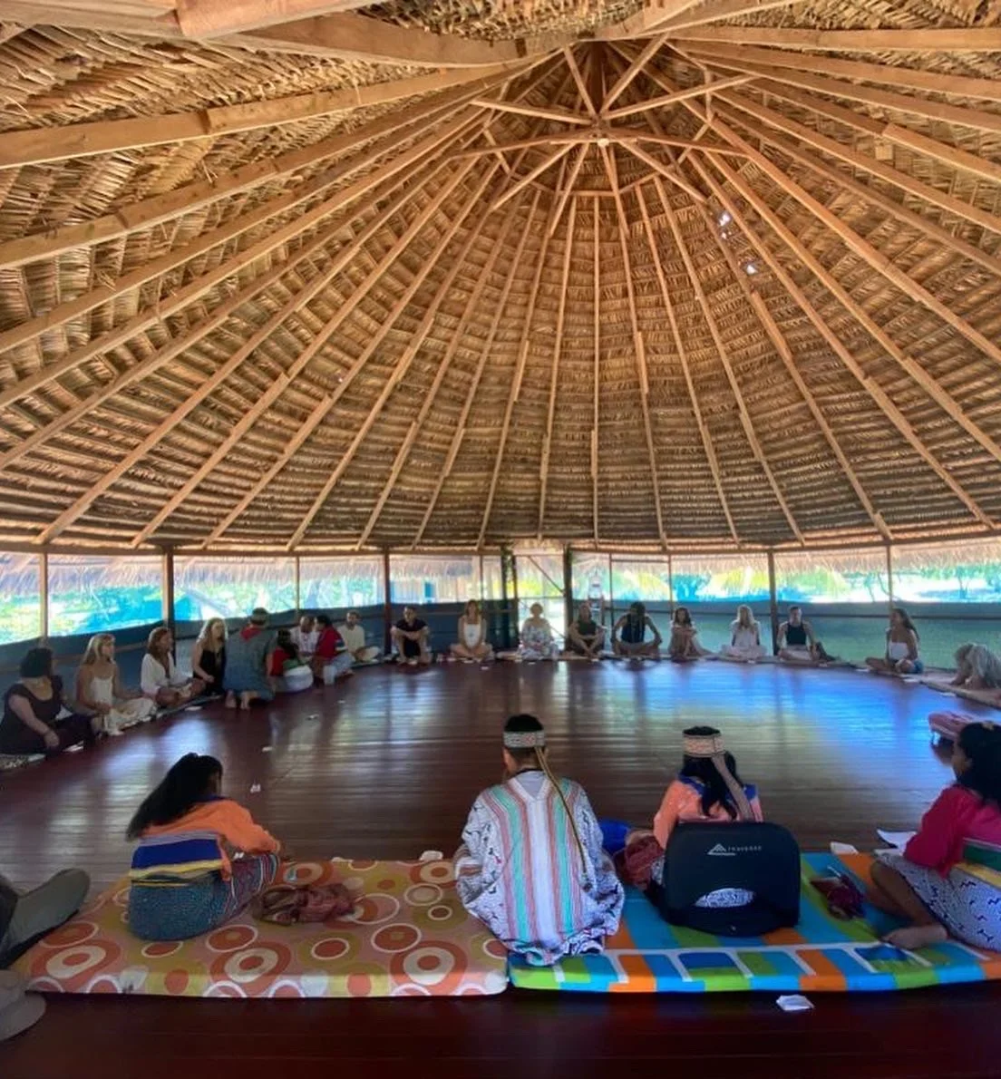 A group of people sitting in a circle inside a large, open, thatched-roof structure, participating in a gathering or ceremony.