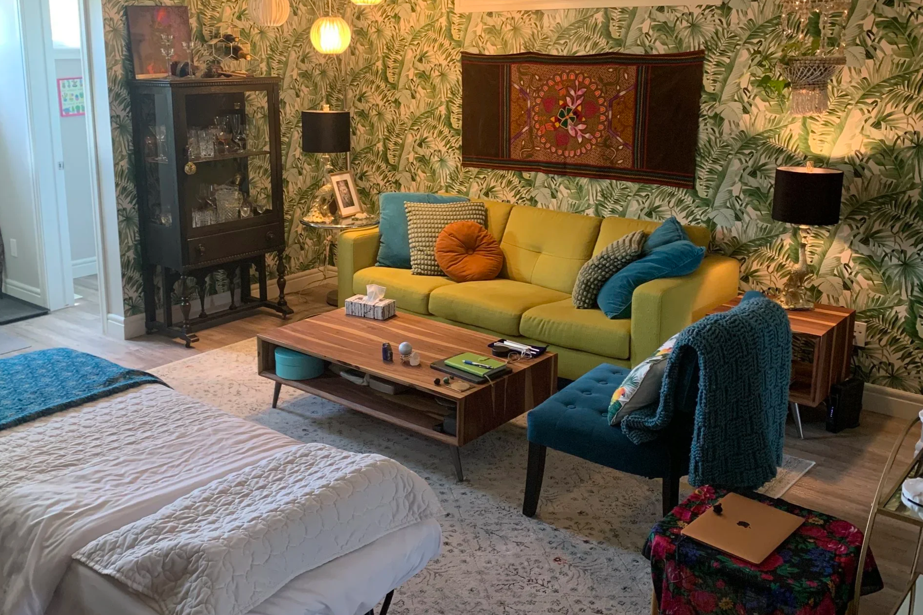 Living room with tropical wallpaper, yellow sofa with multiple pillows, wooden coffee table with miscellaneous items, blue chair with blanket, black display cabinet with glassware, framed photo, illuminated lamps, and wall hanging.