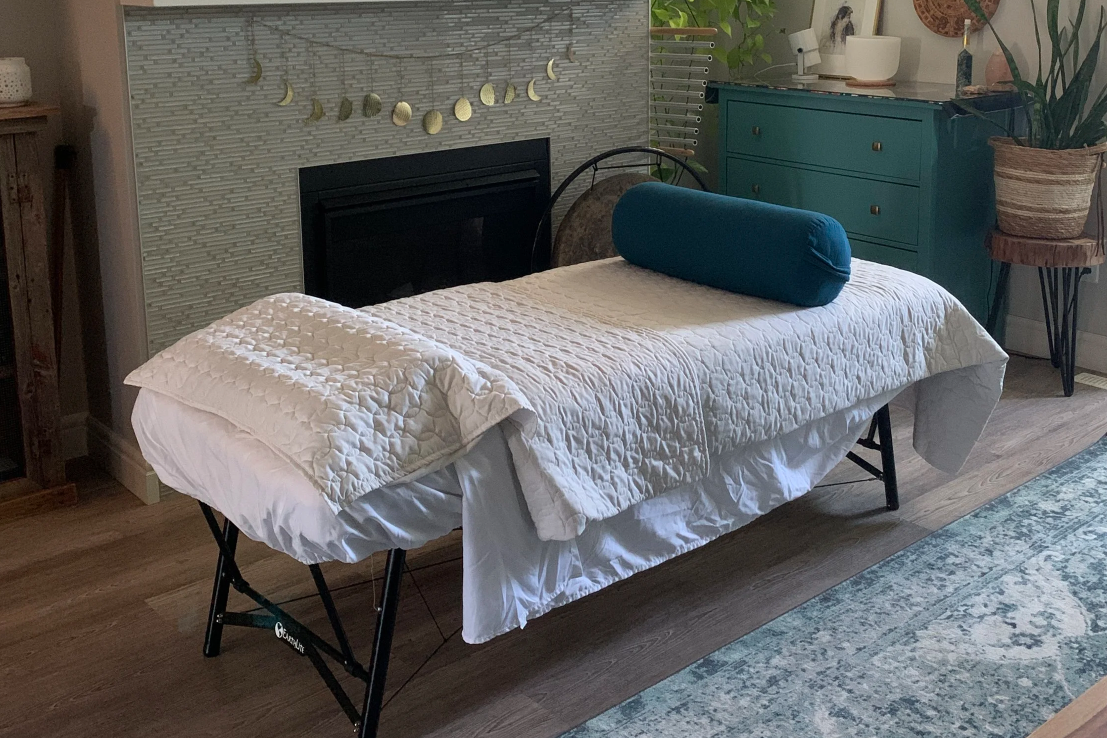A bedroom with a bed covered in a white quilt and a blue cylindrical pillow, a teal dresser on the right, a textured white brick wall behind, and a decorative moon phase wall hanging above the fireplace, with various plants and decorative items around.
