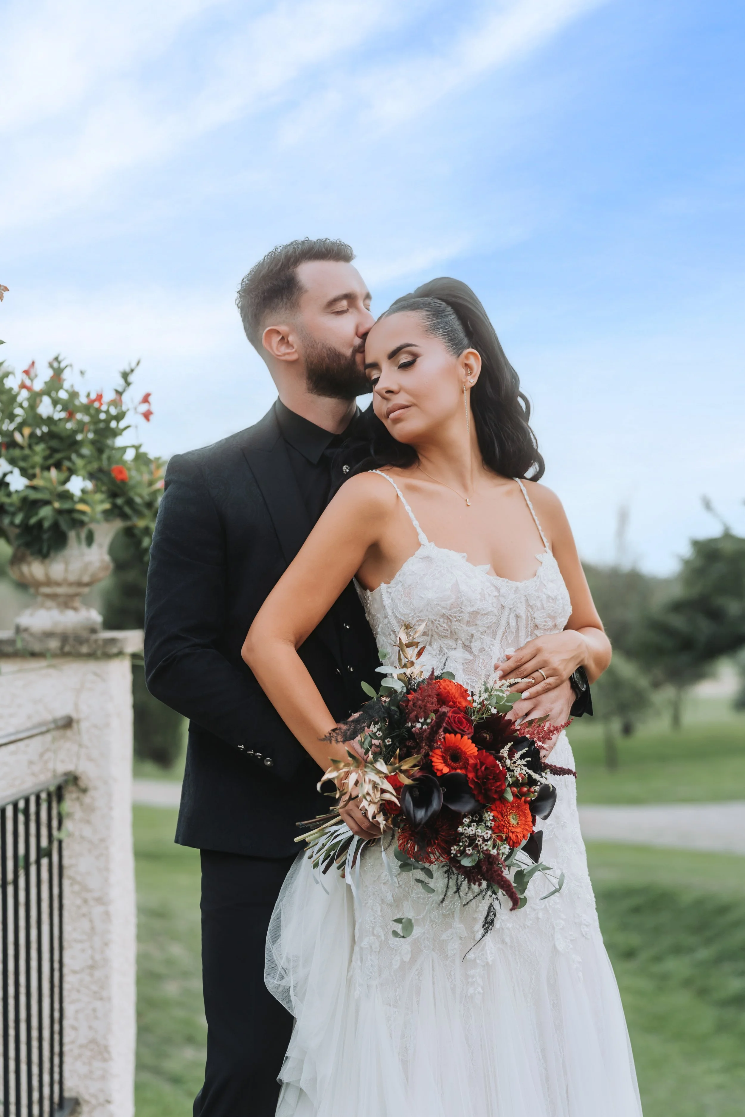 Un couple de mariés en pleine embrassade lors d'une séance photo en extérieur, la mariée tenant un bouquet de fleurs rouges et noires.