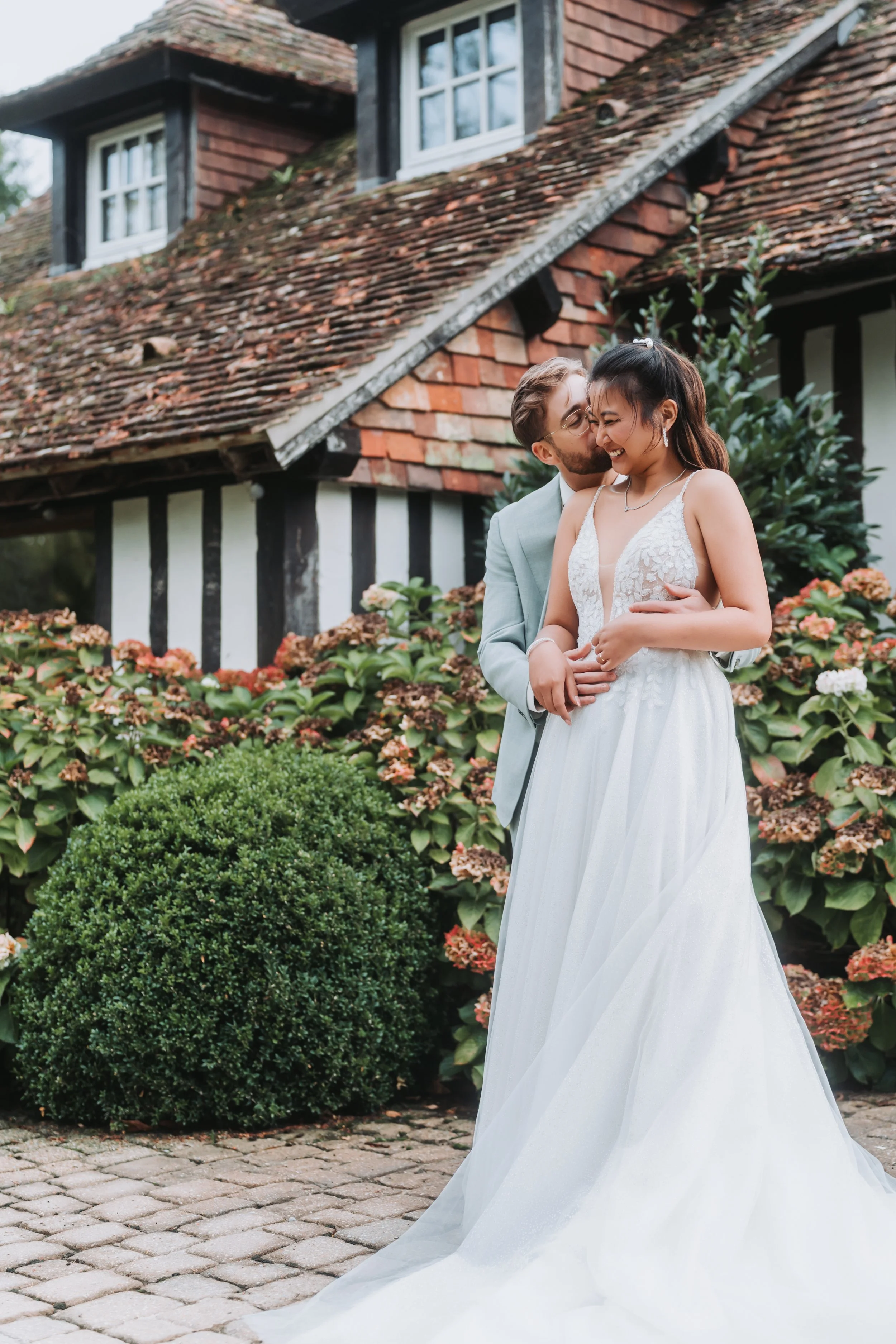 Un couple en habits de mariage s'embrassant et souriant devant une maison en style traditionnel avec un toit en tuiles rouges et des plantes fleuries.