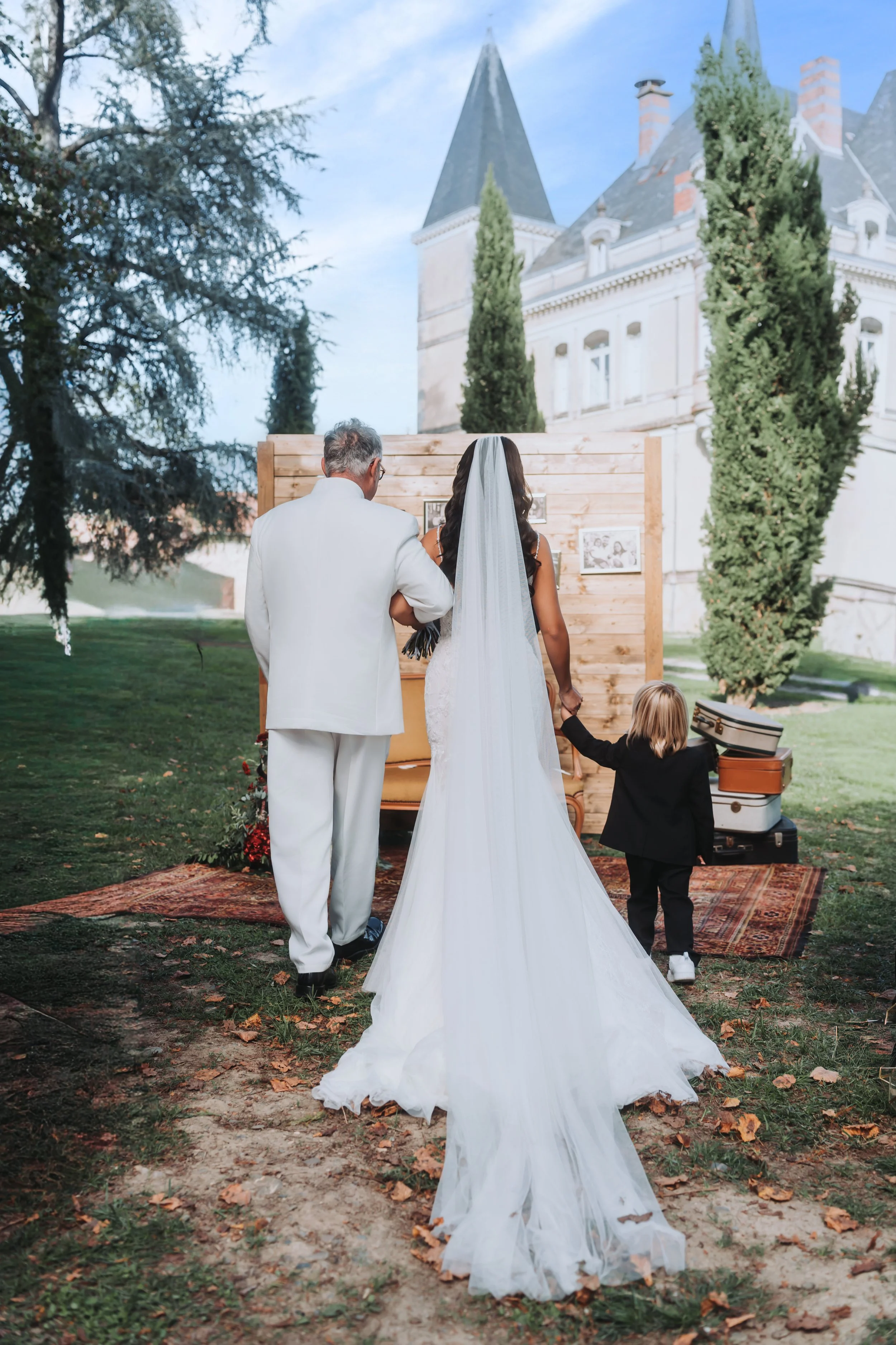 Un mariage en plein air avec une mariée en robe blanche, un homme en costume blanc, une petite enfant en costume noir, un décor vintage avec un tapis, des valises et une cabane en bois, devant un château en arrière-plan.