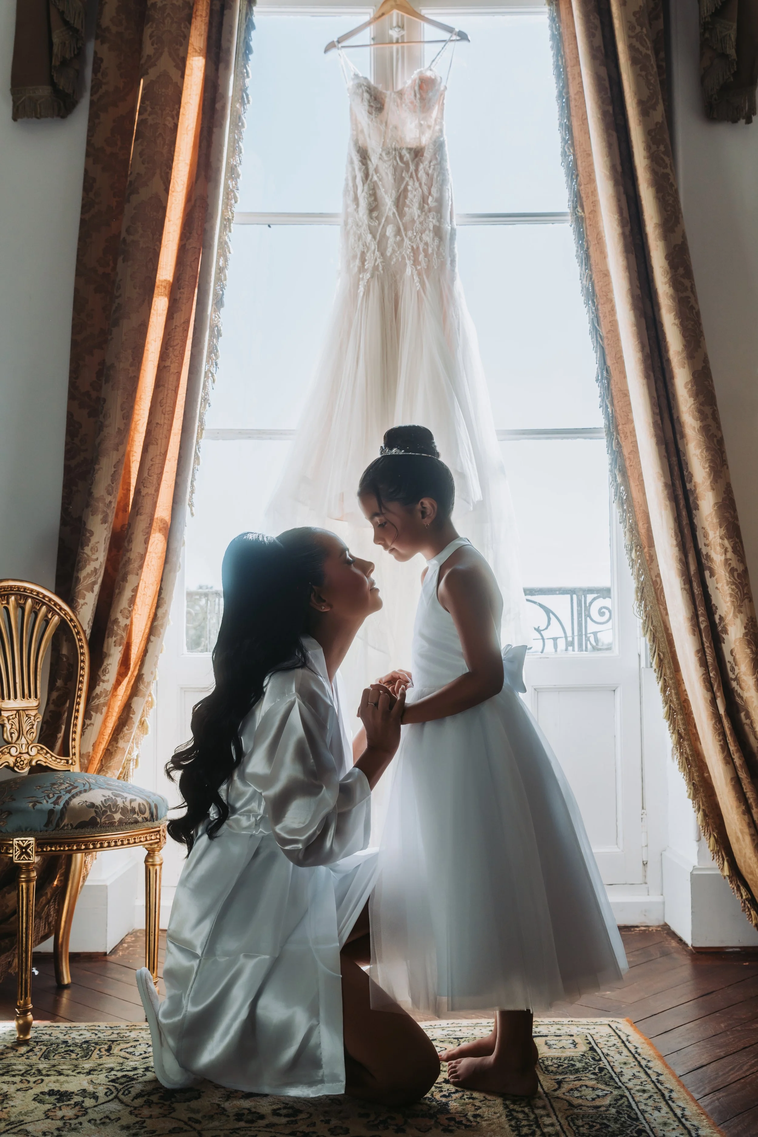 Une femme et une jeune fille en robe blanche se tiennent face à face dans une pièce éclairée par la lumière naturelle d'une fenêtre. Une robe de mariée blanche suspendue derrière elles.