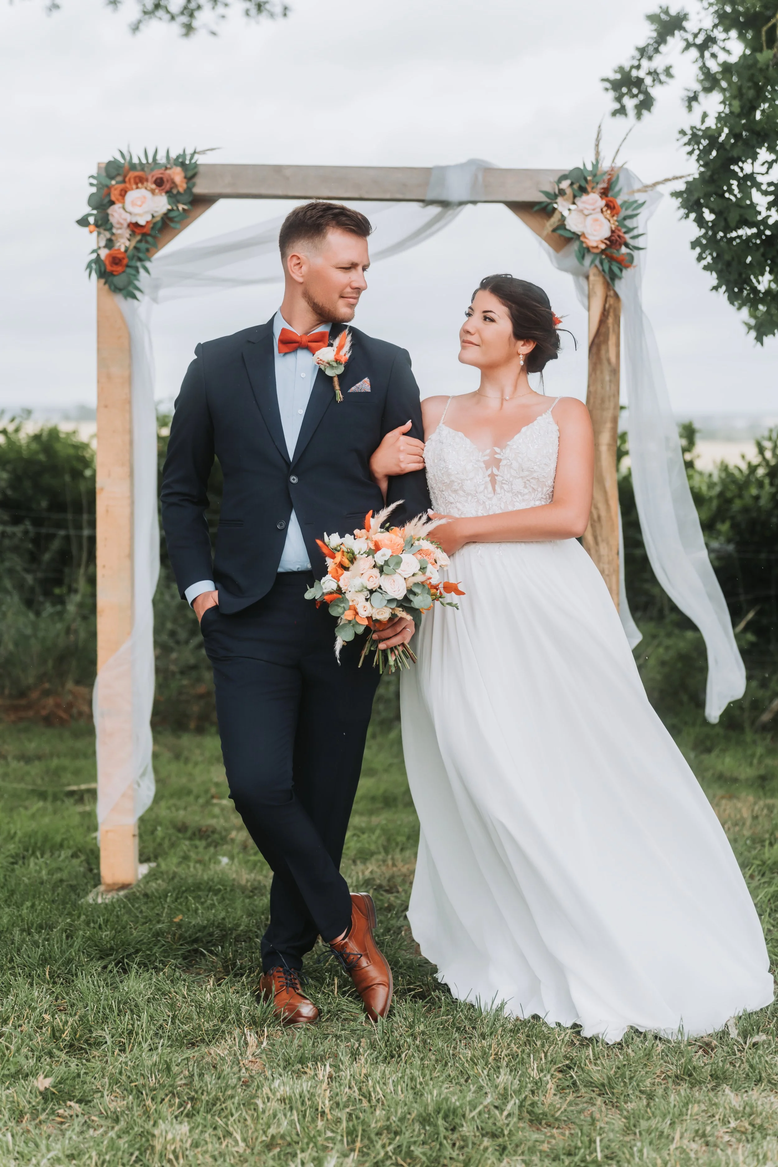 Un couple de mariés marche dans un champ dehors, sous un arc décoré de fleurs. La femme porte une robe blanche et tient un bouquet, l'homme porte un costume sombre avec une cravate orange.