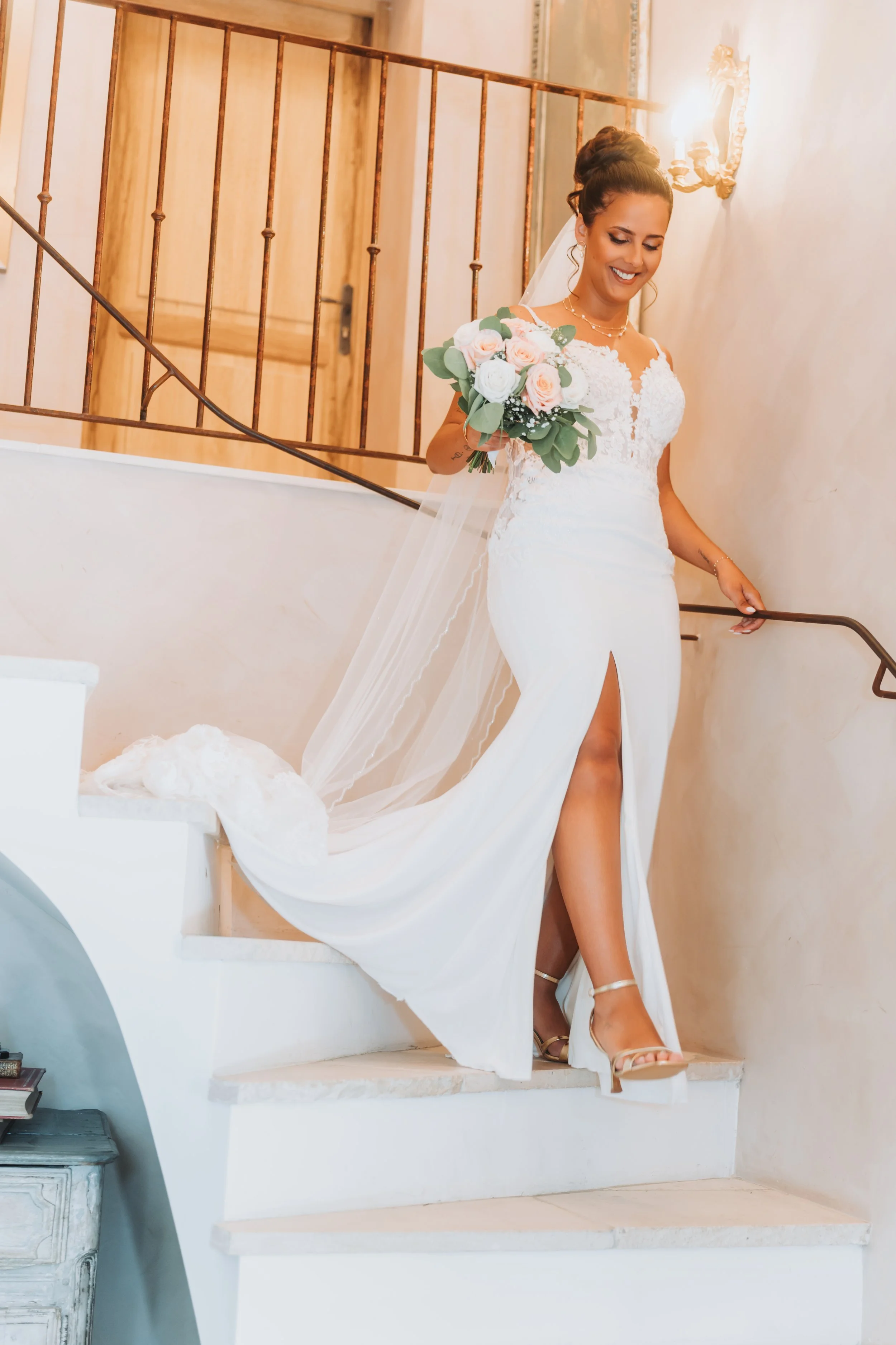 Une femme en robe de mariée blanche descend une escalier en souriant, tenant un bouquet de fleurs roses et blanches.