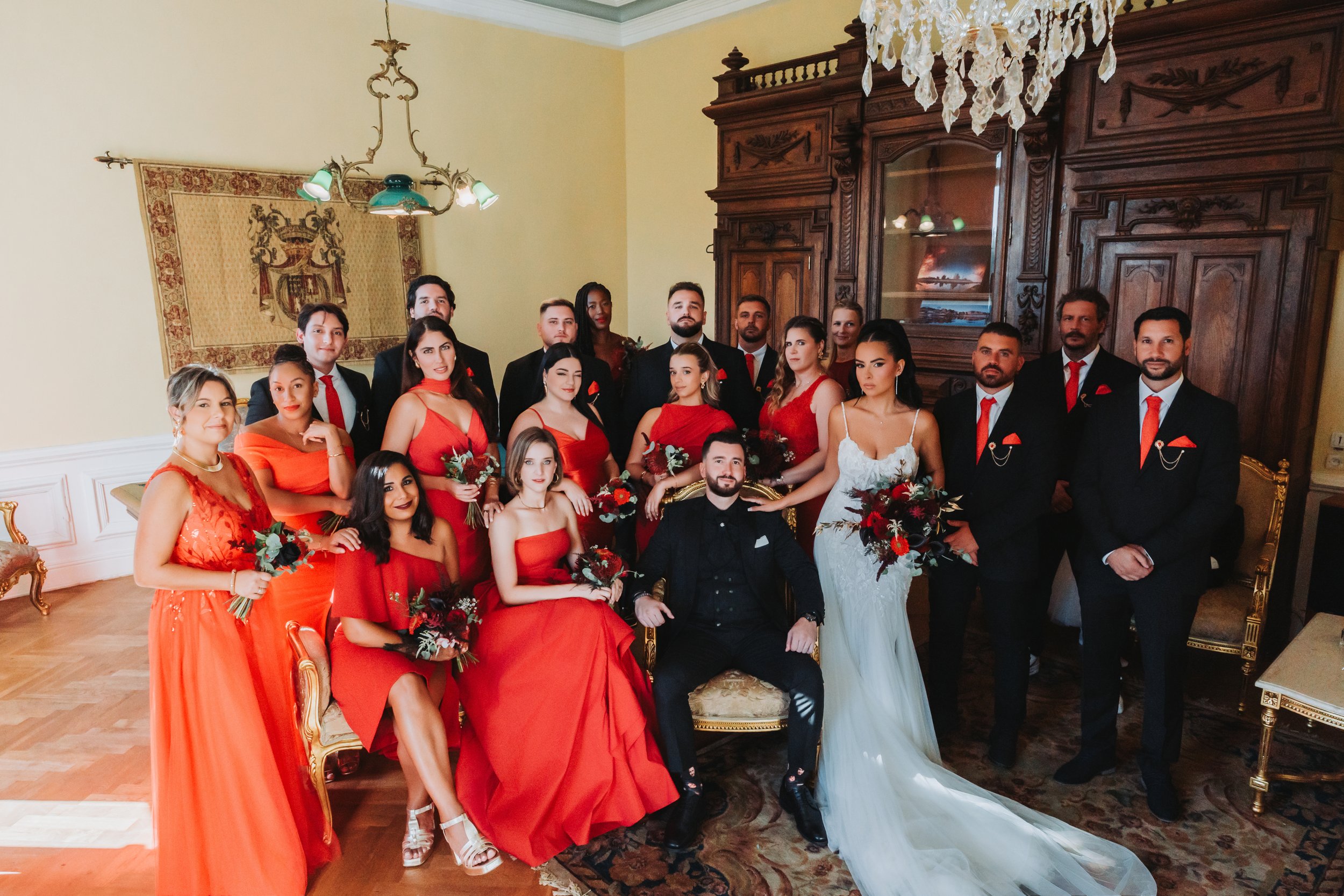 Groupe de personnes lors d'un mariage, avec la mariée en robe blanche et le marié en costume noir, entourés de leurs invités vêtus de rouge et de noir, dans une pièce ornée de bois sculpté et d'un lustre en cristal.