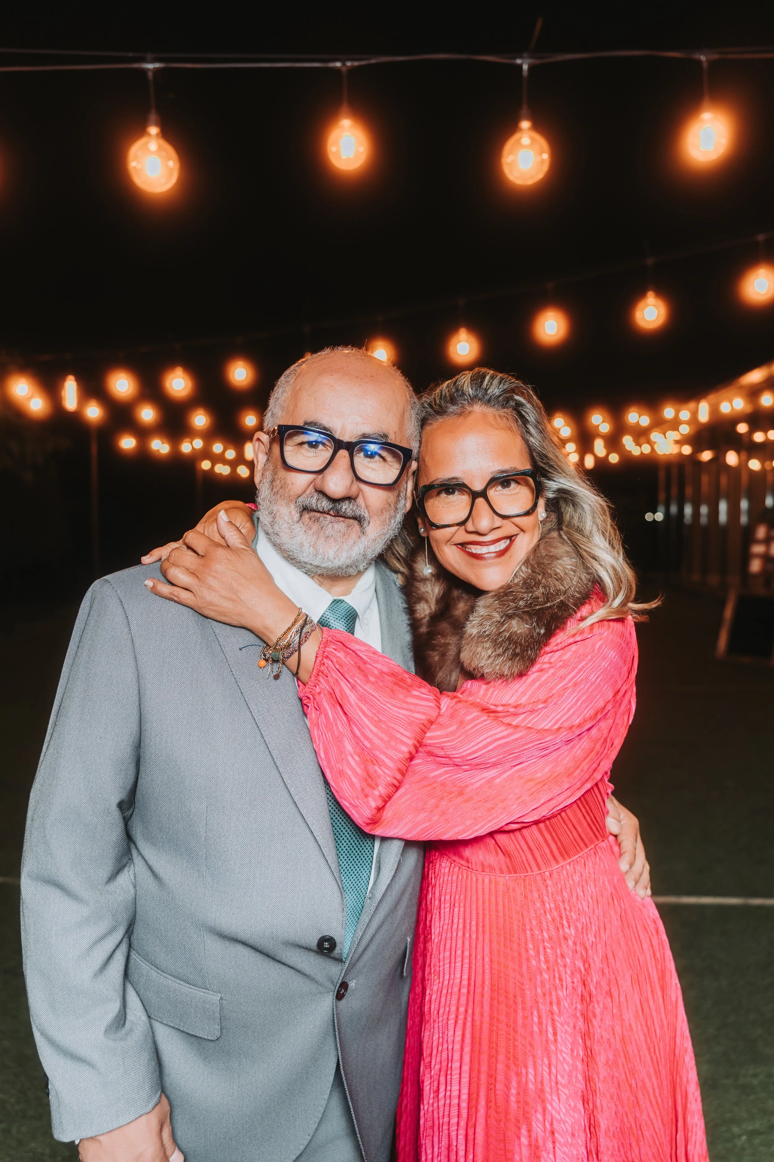 Deux personnes souriantes en soirée sous des guirlandes lumineuses, un homme en costume gris et une femme en robe rouge avec un col en fourrure