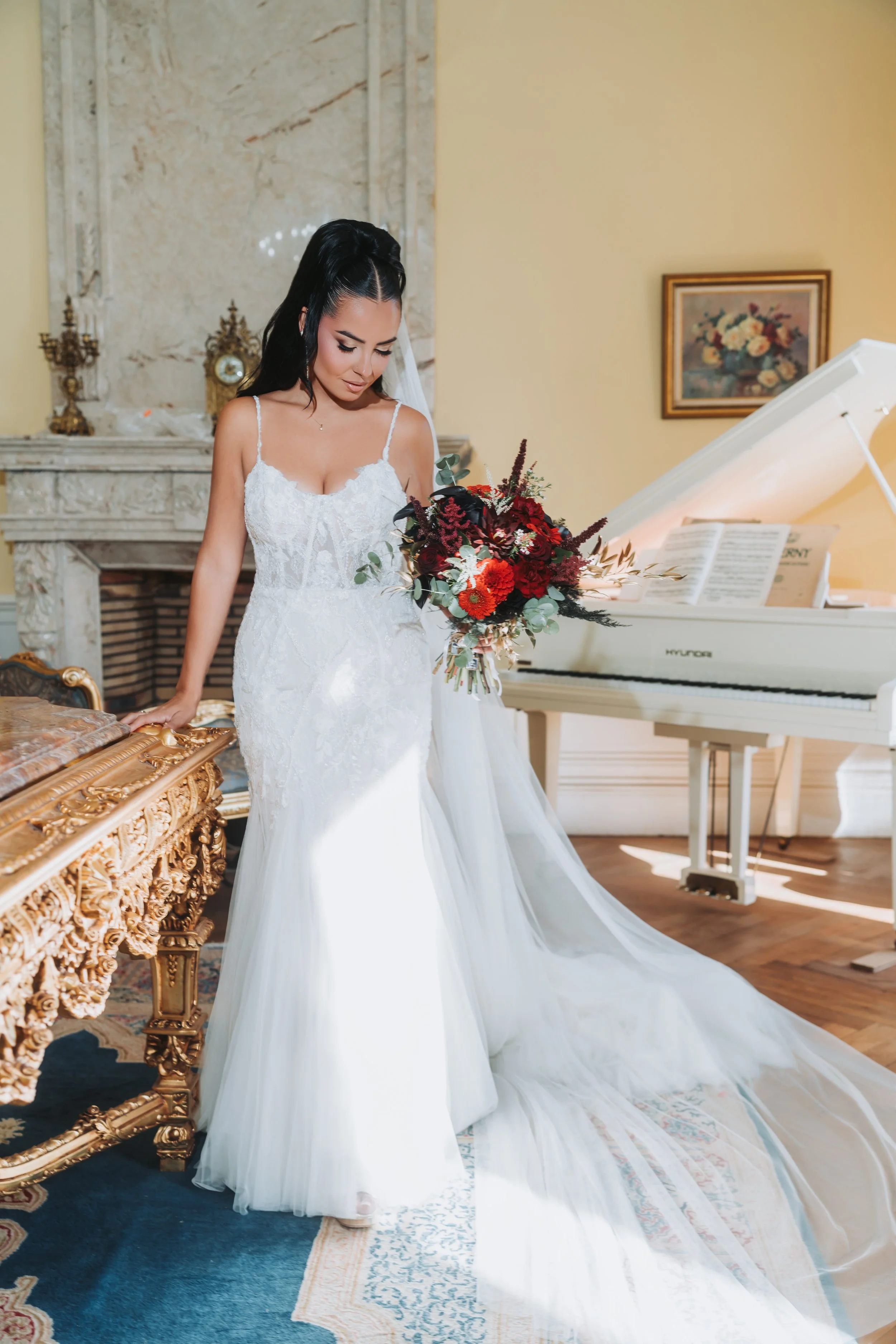 Jeune femme en robe de mariée blanche, tenant un bouquet de fleurs rouges, dans une pièce ornée d'un grand piano blanc et d'une cheminée en marbre.