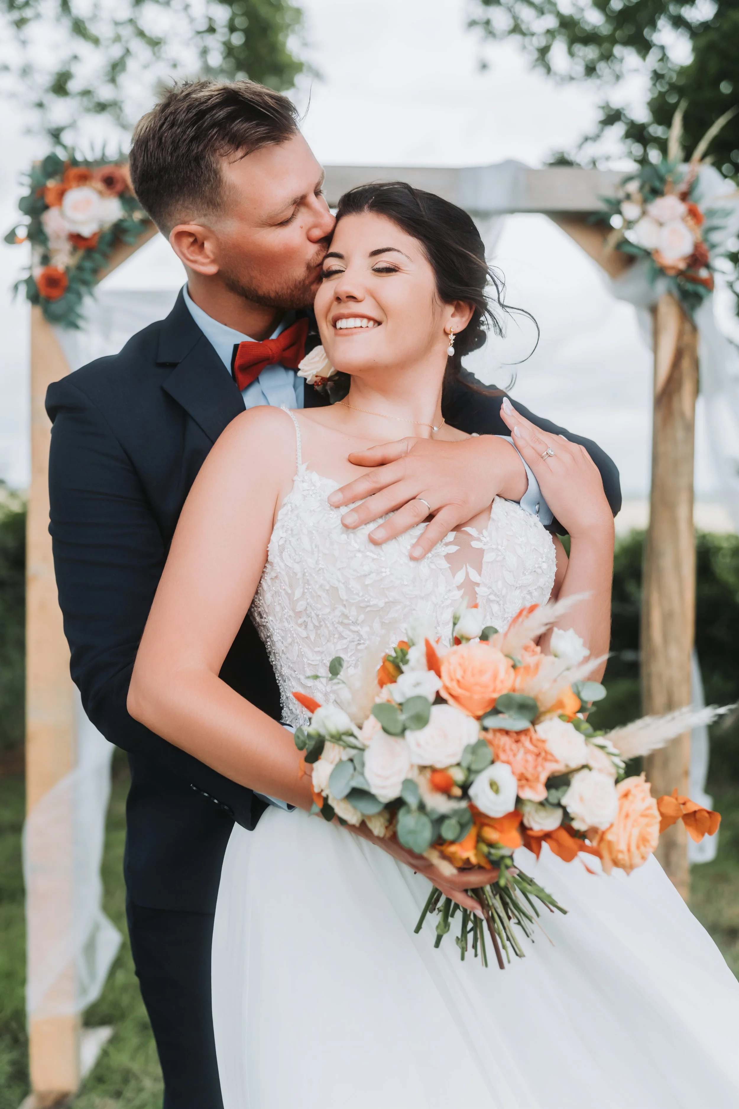 Un couple de mariés en robe de mariée et costume, lors de leur mariage en plein air, devant une arche décorée de fleurs.