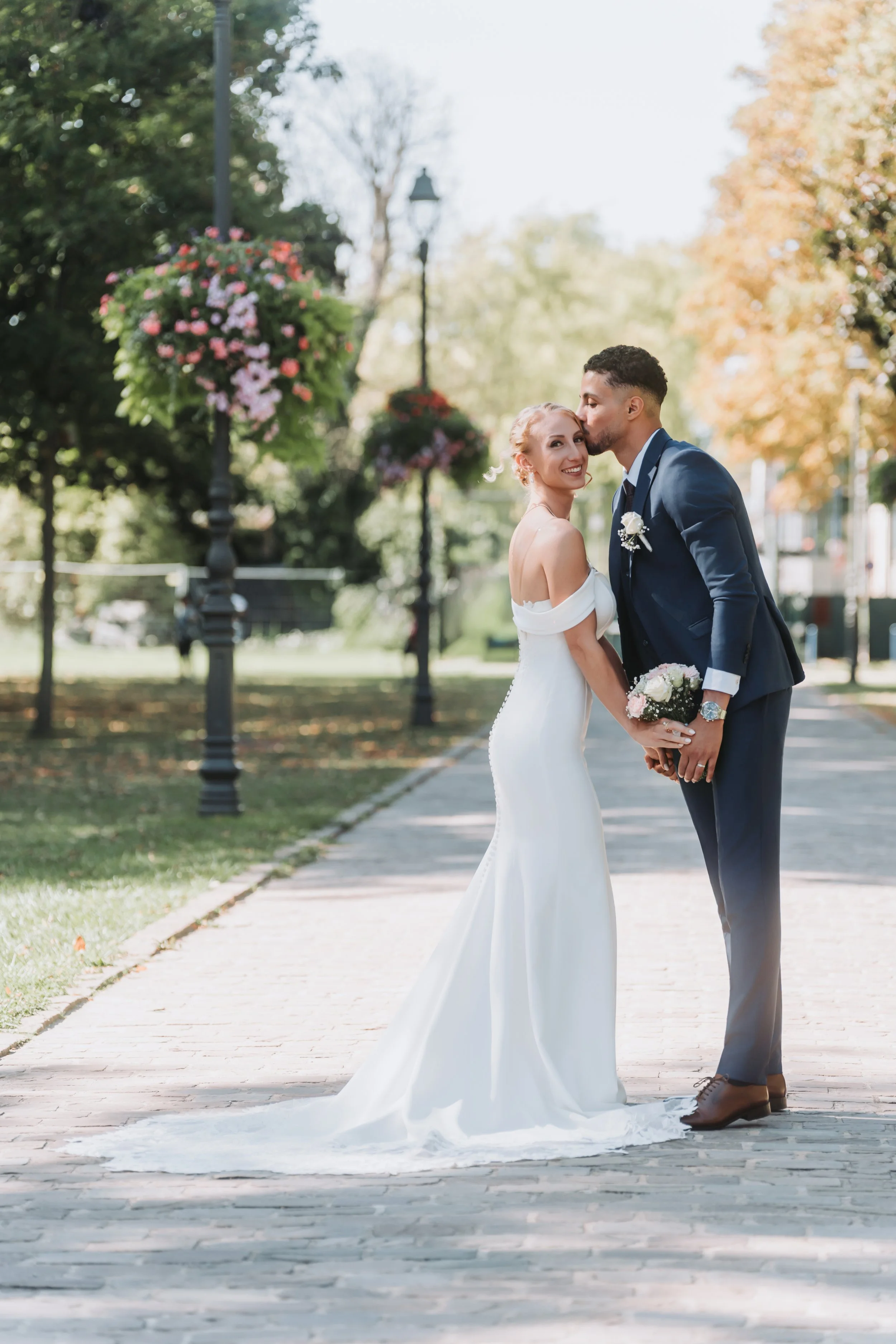 Un couple de mariés en mariage, l'homme embrasse la mariée sur le front dans un parc, avec des arbres, des lampadaires et des fleurs en arrière-plan.
