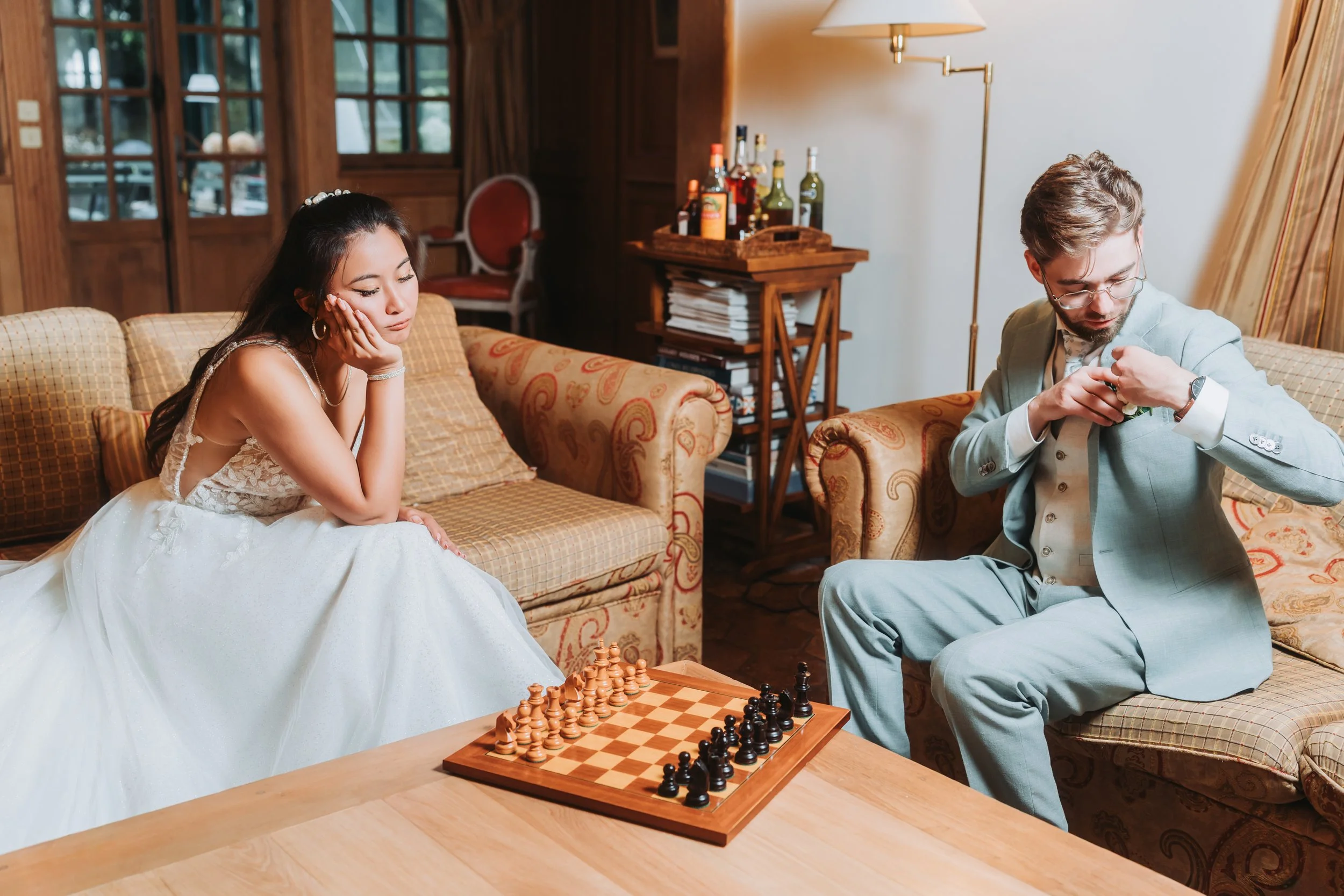 Une femme en robe de mariée assise sur un canapé, semblant pensif, et un homme en costume qui regarde sa montre dans un salon élégant.