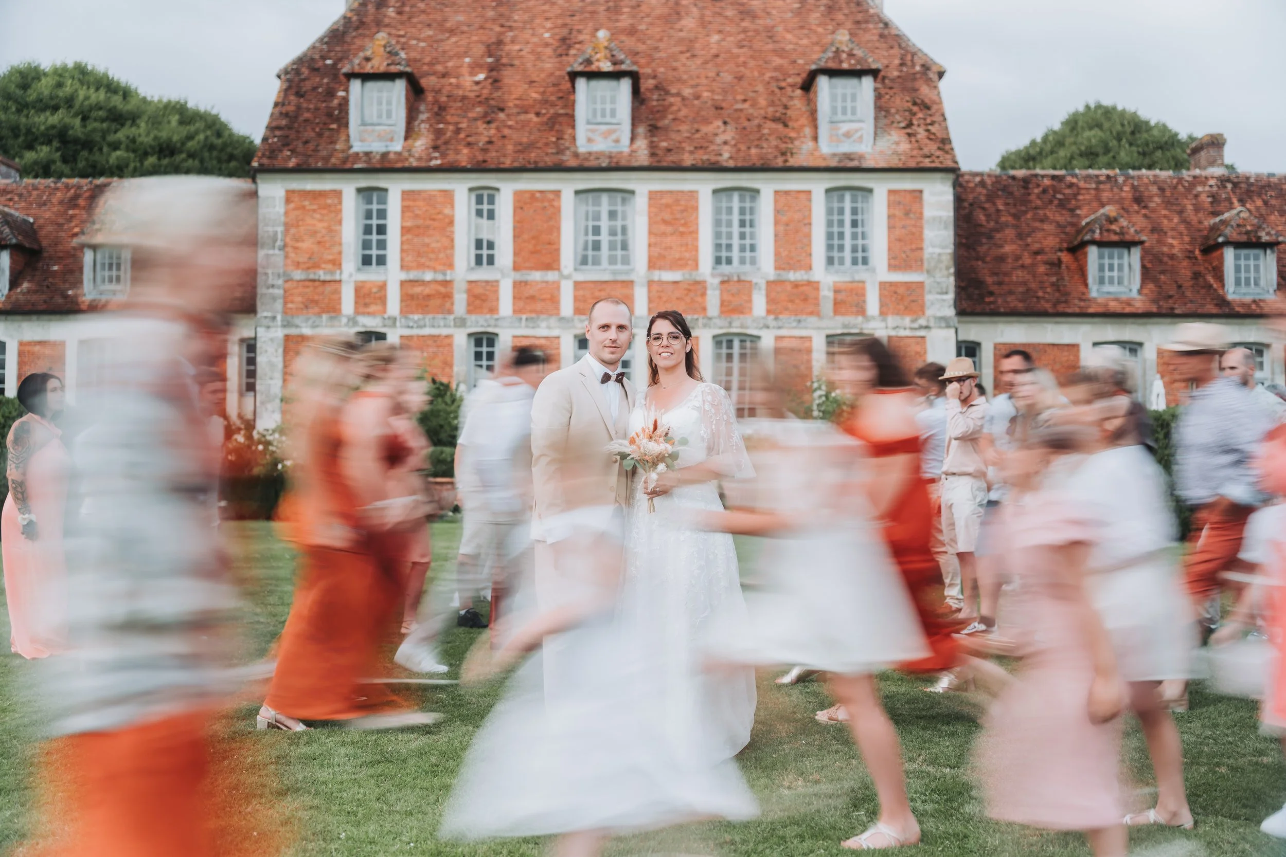 Un couple de mariés posant devant un bâtiment ancien, entourés de personnes floues qui dansent lors d'une fête de mariage en extérieur.