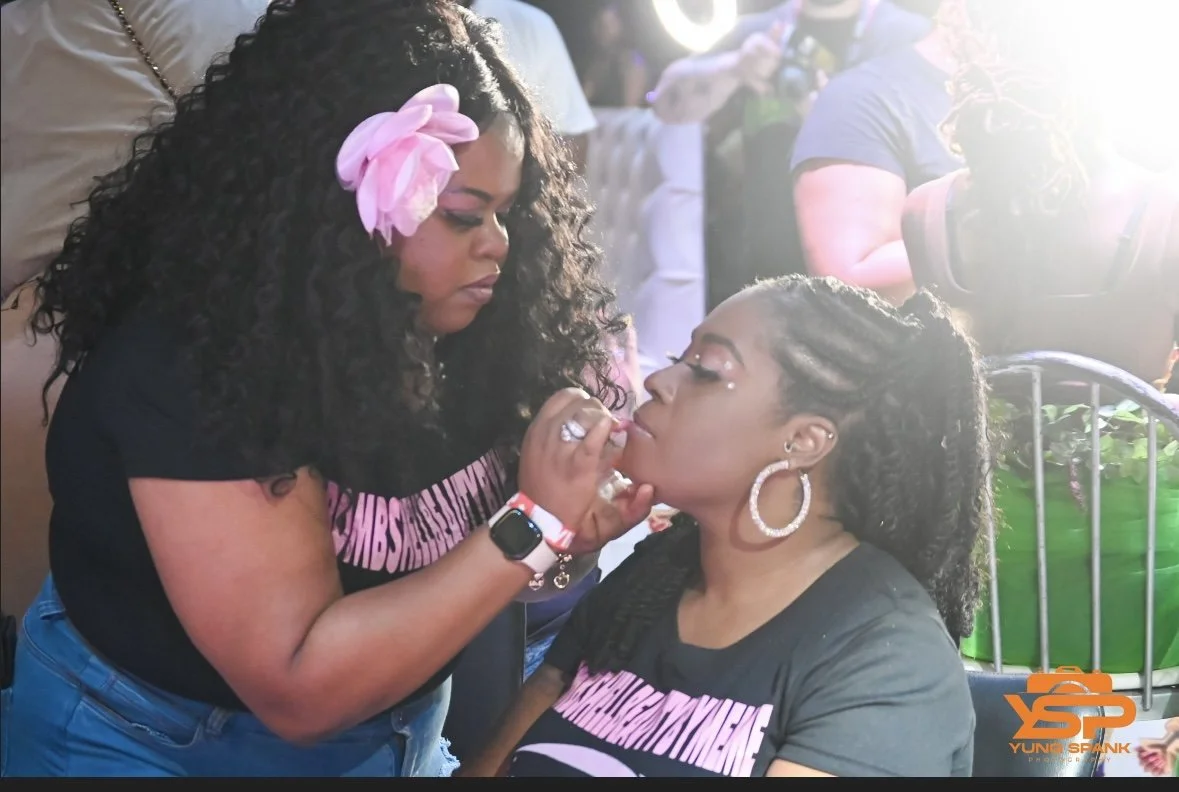 A woman with curly hair and a pink flower in it applying makeup or lipstick to another woman with braids and hoop earrings at a crowded outdoor event.