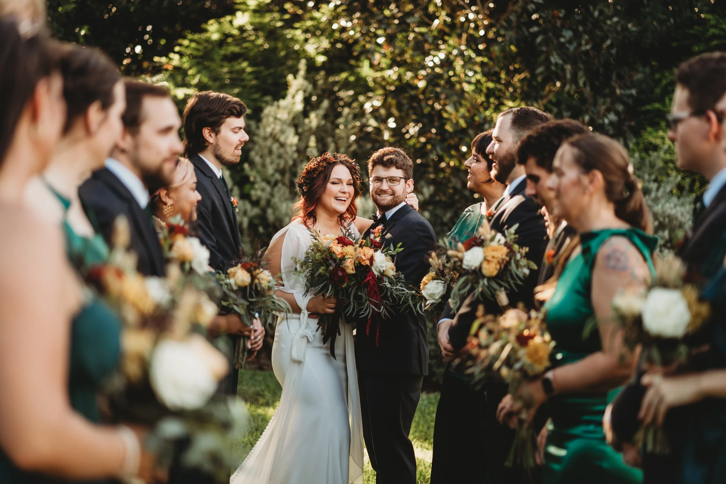 Bride and groom embracing amidst wedding party holding bouquets outdoors at sunset.