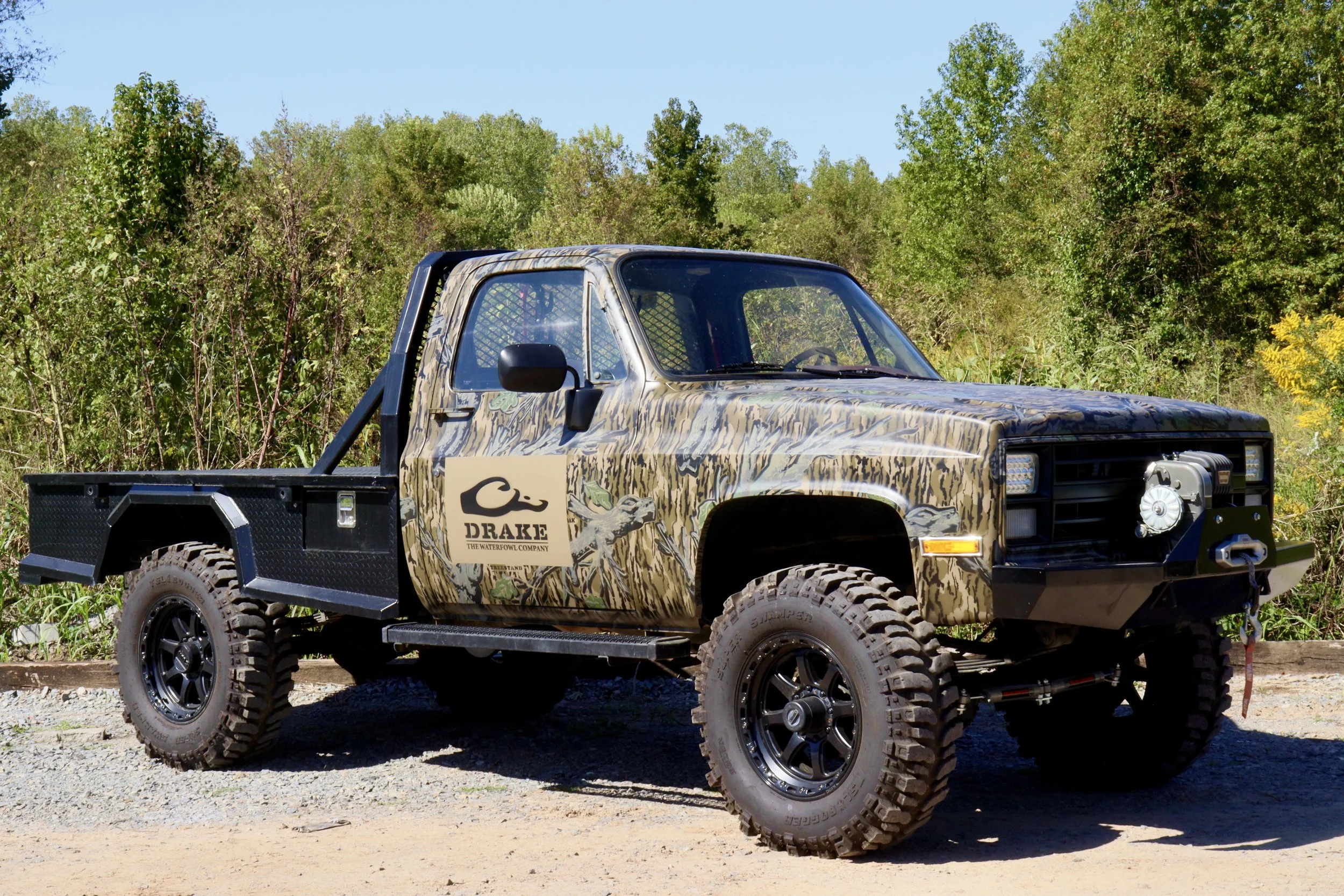 A camouflaged off-road truck with a logo on the door that says Drake, parked outdoors on dirt with green bushes and trees in the background.