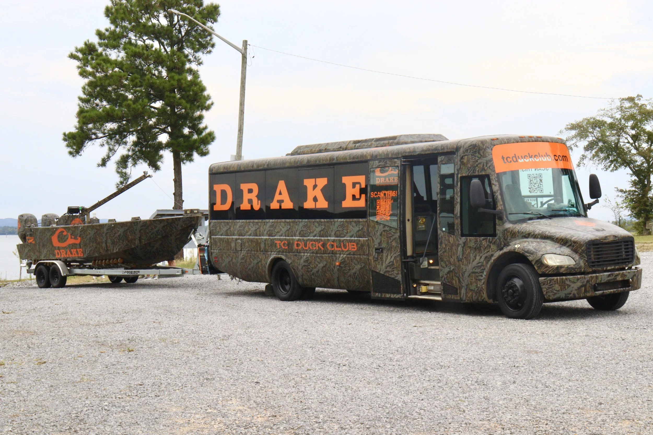 Camouflage-patterned bus with orange lettering reading 'DRAKE' and 'TC DUCK CLUB', attached to a boat with the same branding, parked on gravel near trees and a body of water.