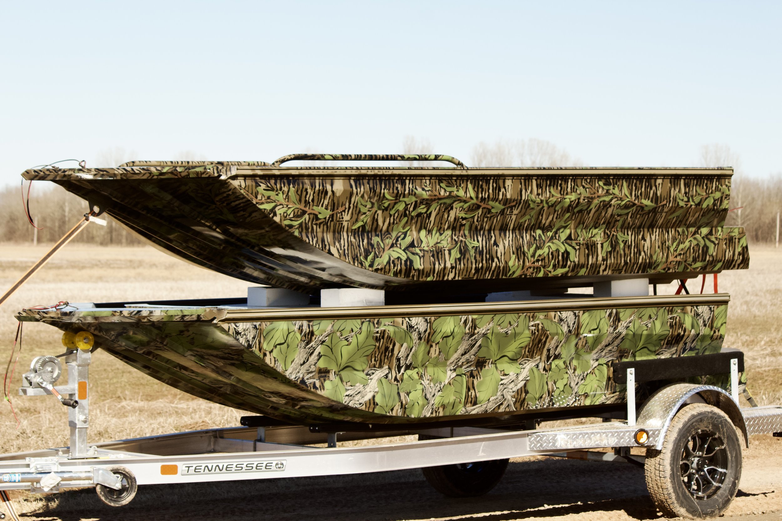 A person kneeling under a boat in a garage or workshop, holding a power drill, wearing a mask and gloves, with tires and chains nearby.