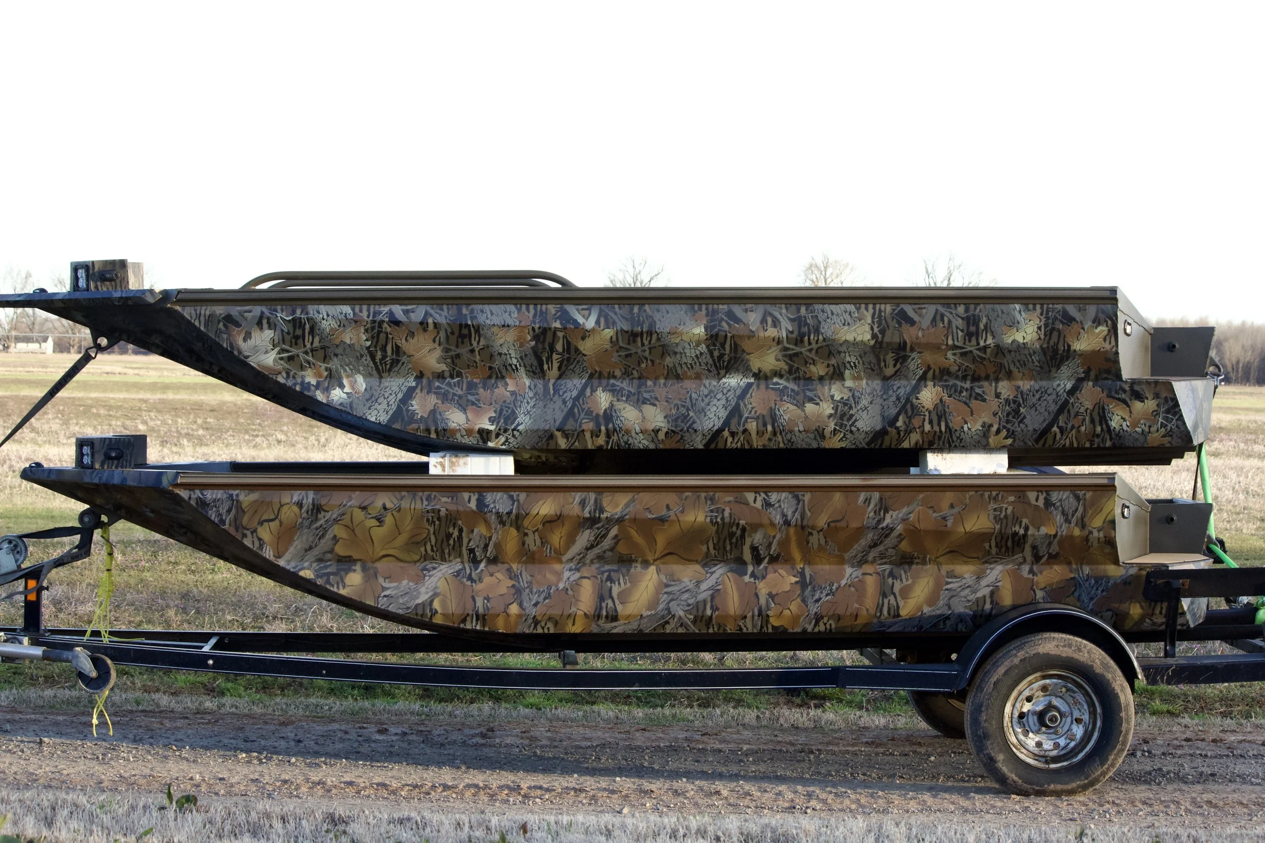Two camouflage-patterned boats on a trailer in a field.