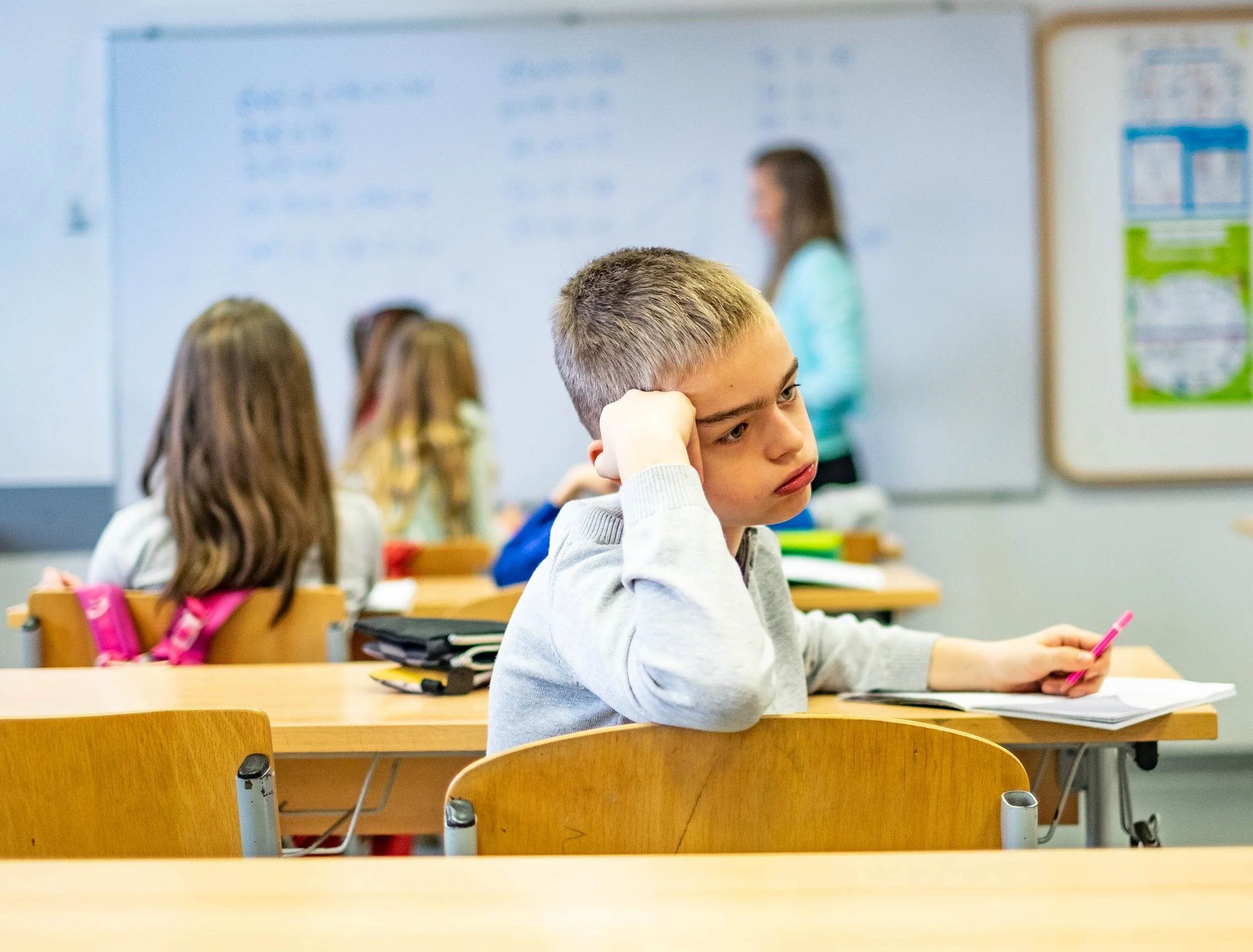 A young boy sitting at a classroom desk, resting his head on his hand, looking thoughtful, with a pink pen and notebook in front of him. Other students are visible in the background, and a teacher is at the whiteboard.
