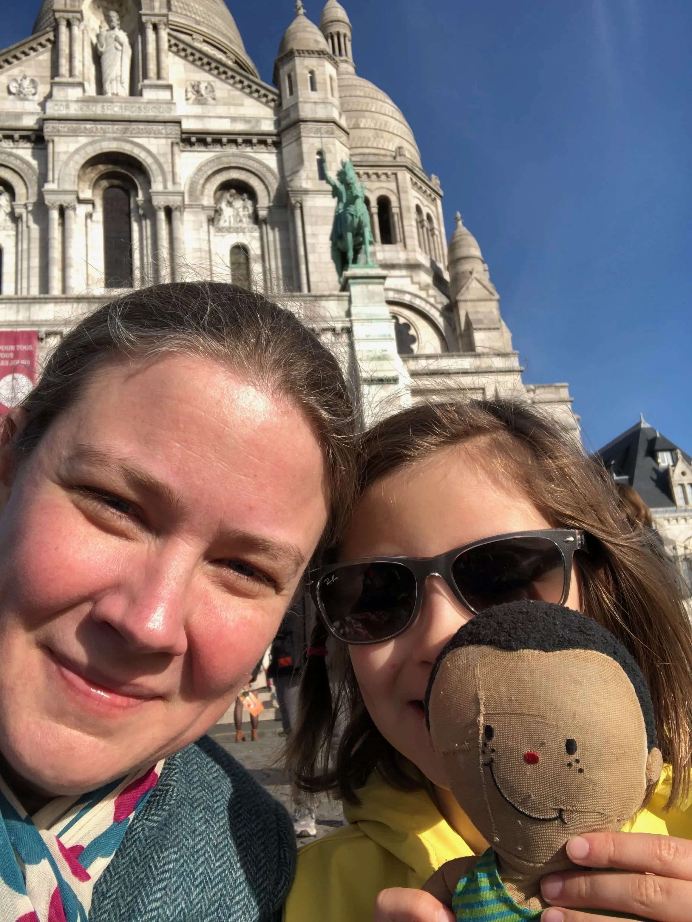 Two women taking a selfie in front of a historic European-style building with domes and a statue of a horse and rider. One woman is smiling, while the other is holding a plush doll with a smiling face and wearing sunglasses.
