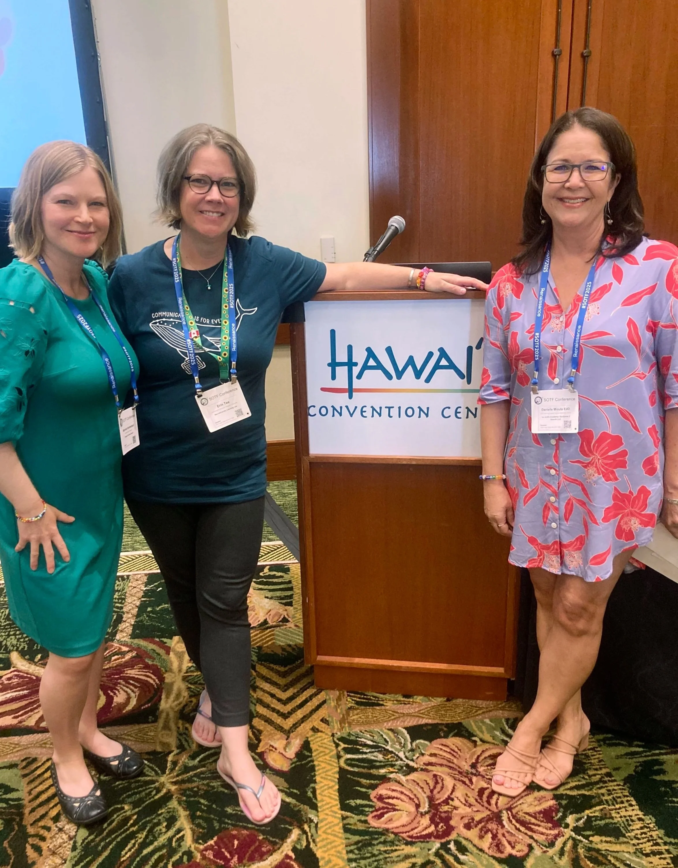 Three women standing next to a sign that reads 'Hawaii Convention Center'. They are attending a conference, indicated by their name badges and lanyards. They are smiling and posing for the photo.