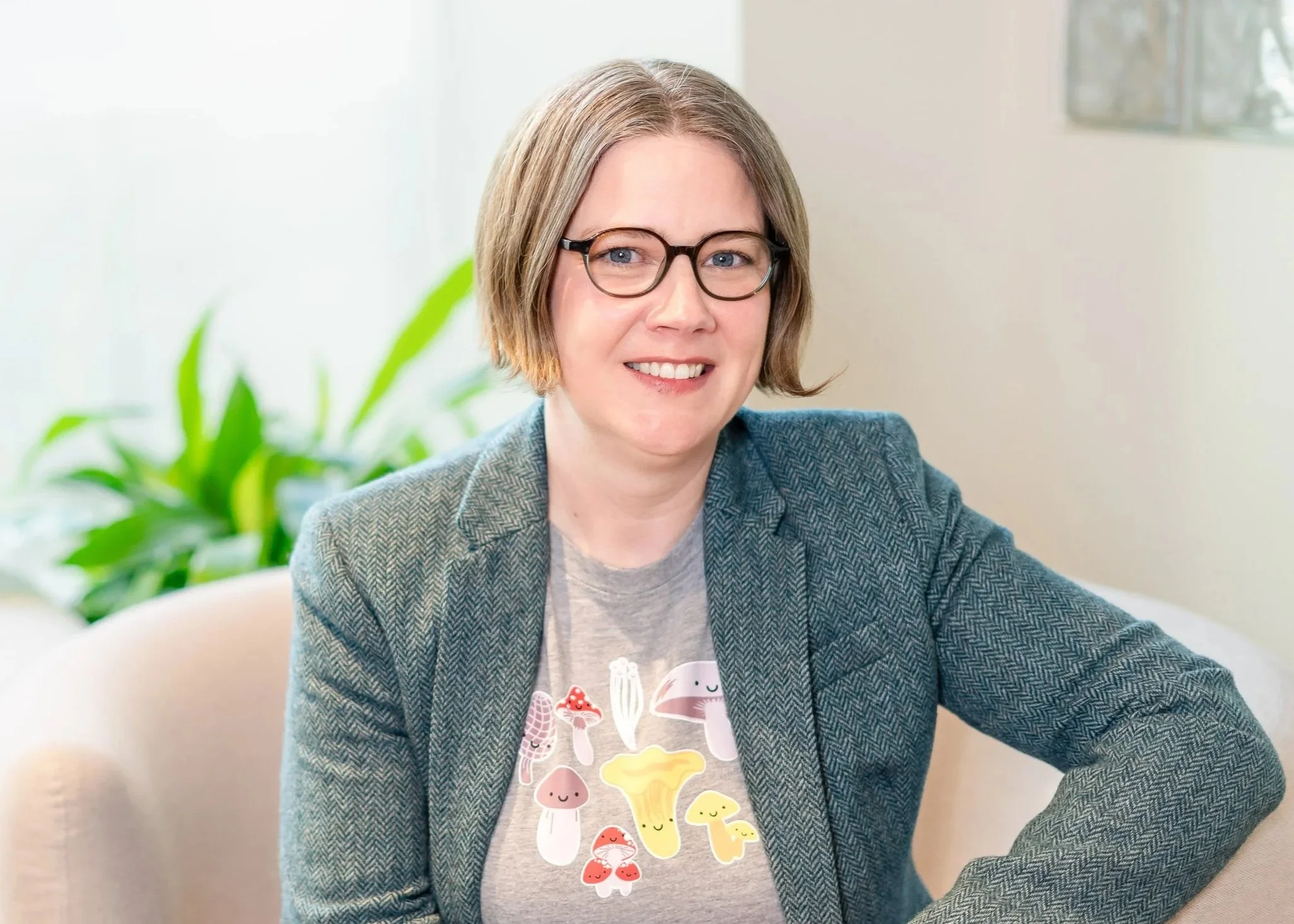 A woman with short blonde hair, glasses, and a grey t-shirt with colorful mushroom illustrations, sitting on a beige armchair in a bright room with a plant and window in the background.