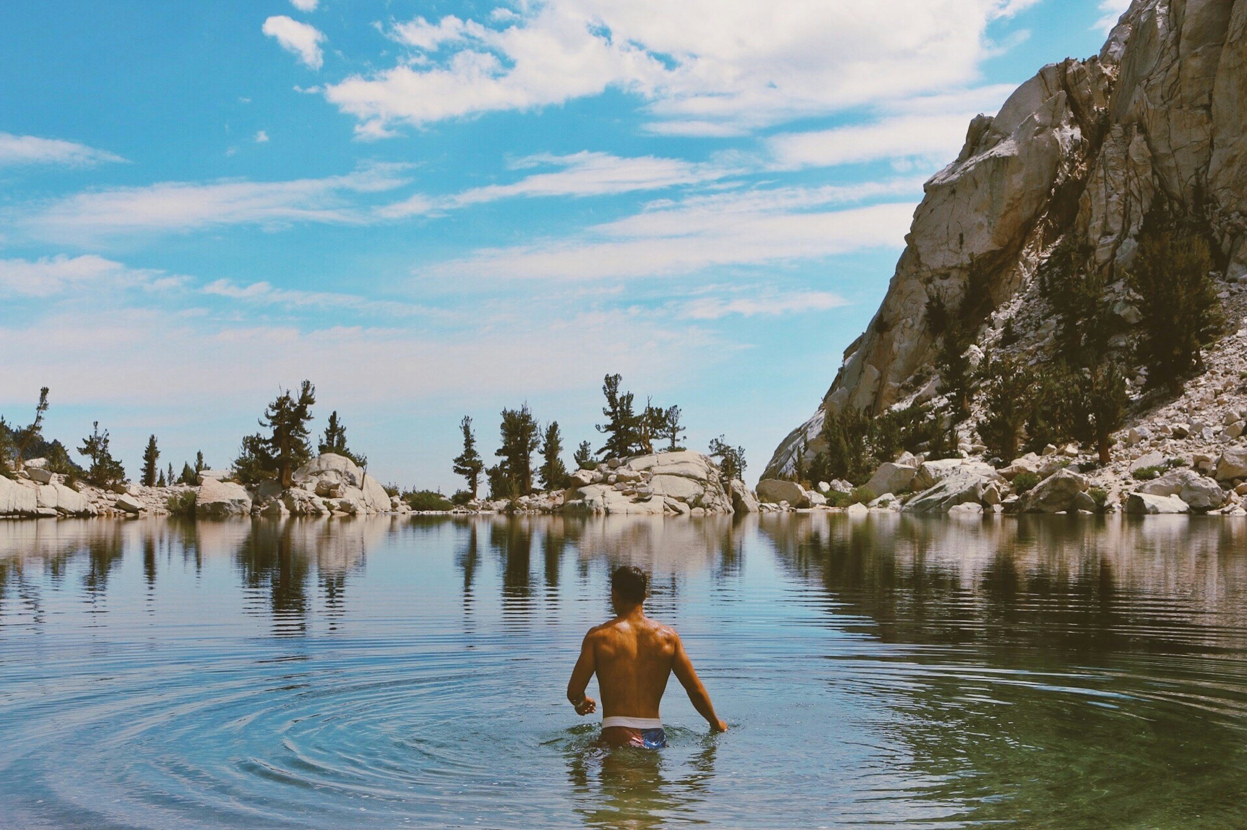 A man in swimming trunks wading into a calm mountain lake, surrounded by rocky cliffs and pine trees under a blue sky with scattered clouds.
