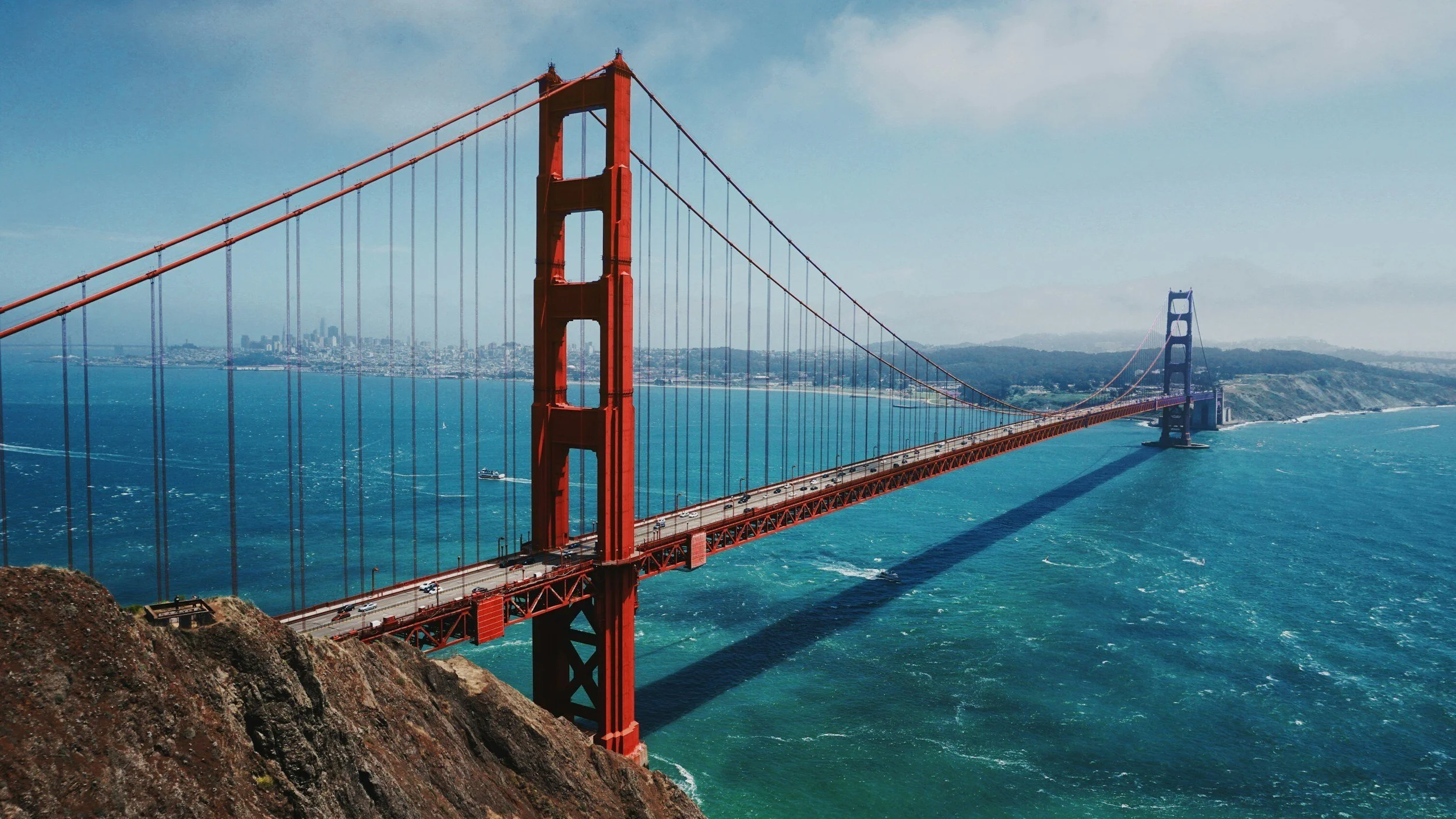 Aerial view of the Golden Gate Bridge spanning the San Francisco Bay with city skyline in the background, clear blue water below, and partly cloudy sky.