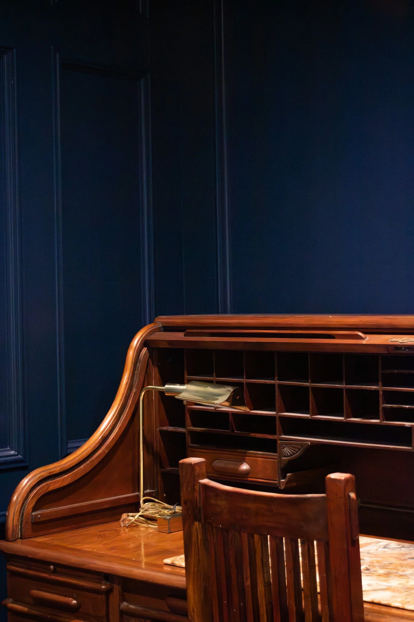 A wooden desk with a sloped top and multiple small cubbyholes, with a brass desk lamp and a wooden chair placed in front of it, against dark blue paneled walls.