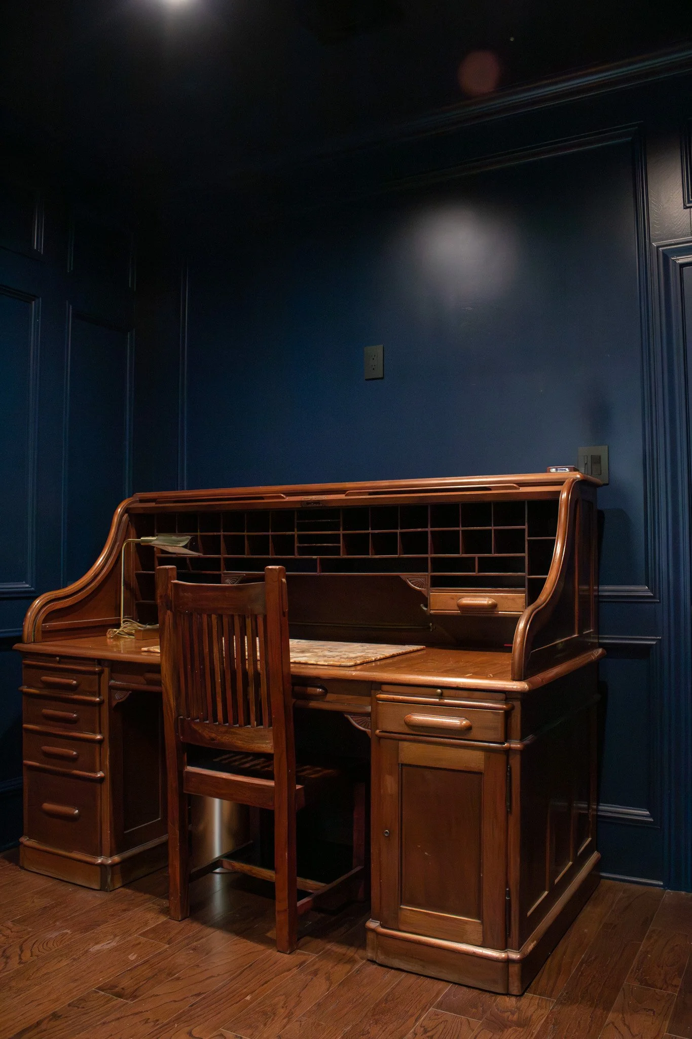 A vintage wooden writing desk with a chair in front of it, set against dark blue paneled walls and wooden flooring.