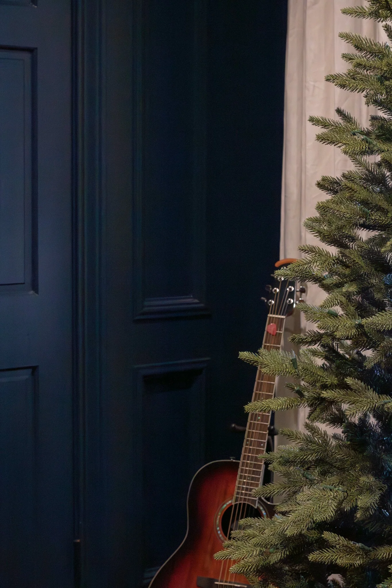 Partially visible Christmas tree on the right and an acoustic guitar leaning against a dark wall with paneling on the left.