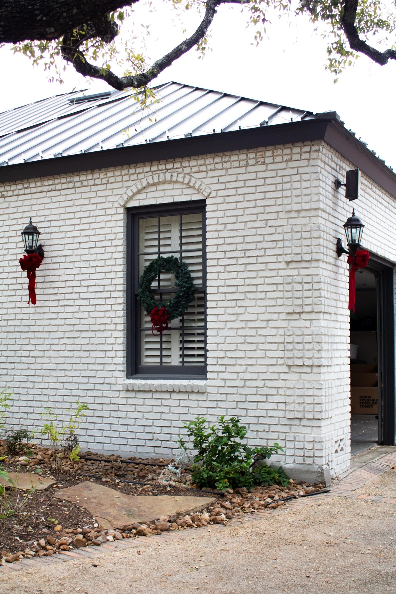 White brick house decorated for Christmas with a wreath on the window, two wall-mounted lanterns with red ribbons, and a small garden with plants and a stone pathway in front.