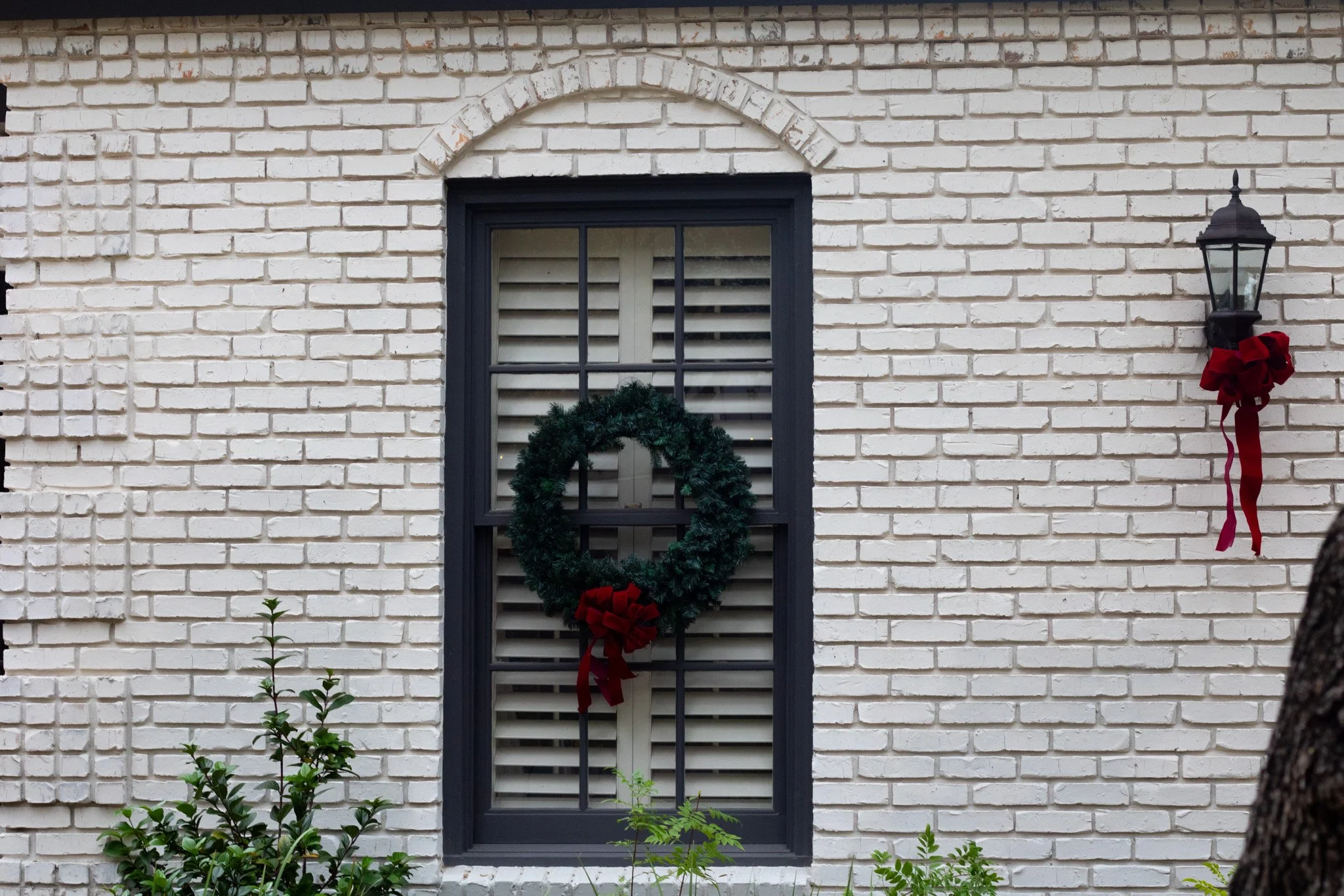 White brick house exterior with black-framed window decorated with a green Christmas wreath with red bow, and a black lantern style outdoor light with a red bow on the right side of the window.