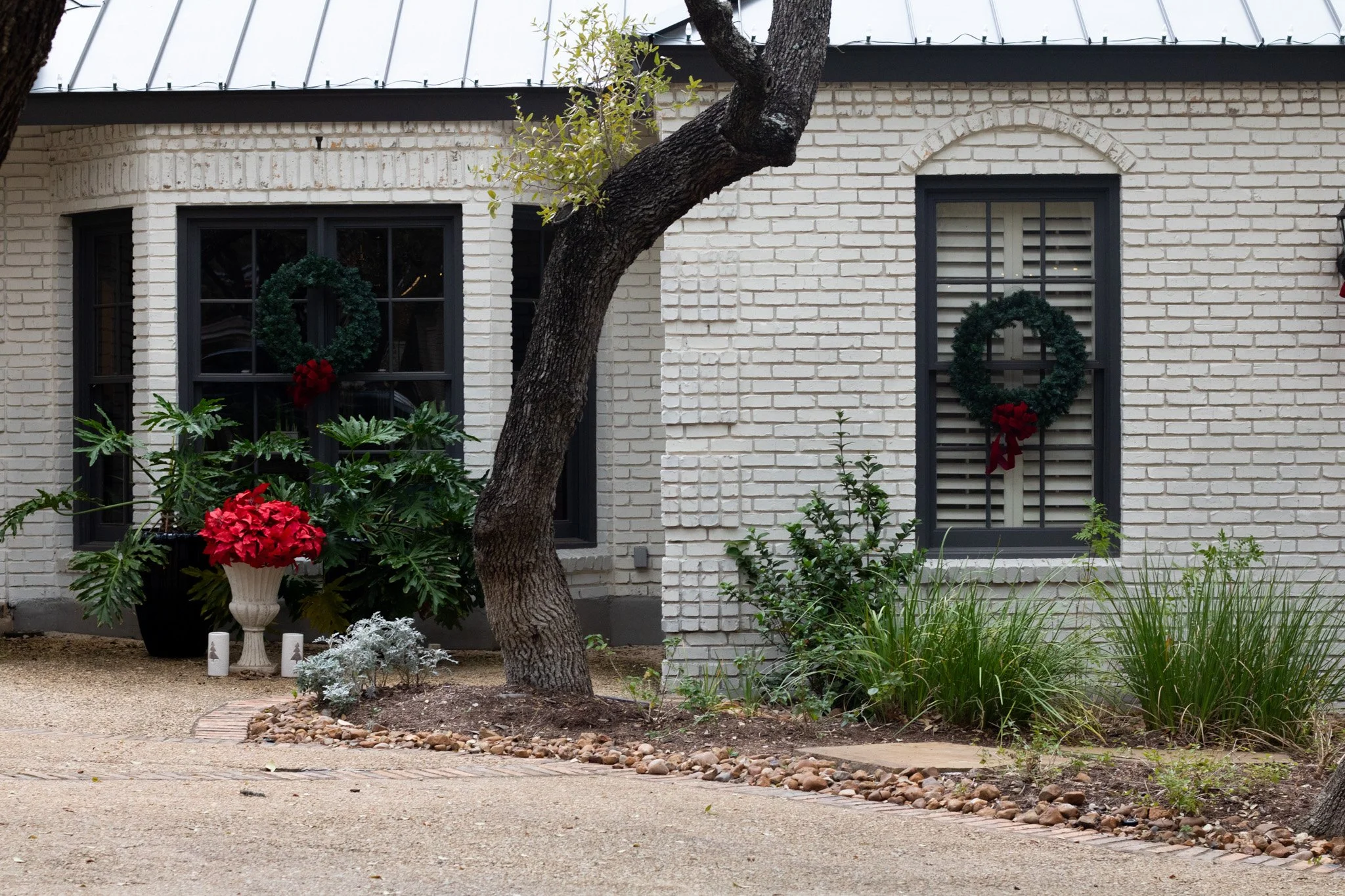 A white brick house with black window frames decorated with Christmas wreaths. There are potted plants and bushes outside, and a curved pebble border around a small garden bed.