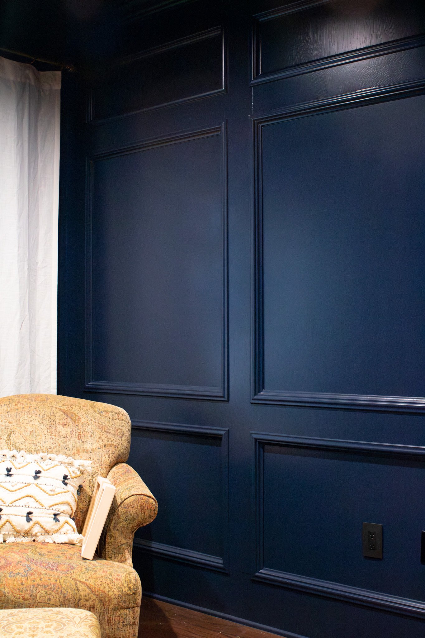 A cozy corner with a blue paneled wall, part of an upholstered armchair with patterned fabric, a folded blanket on the armrest, a closed book, white curtains, and a dark wooden floor.