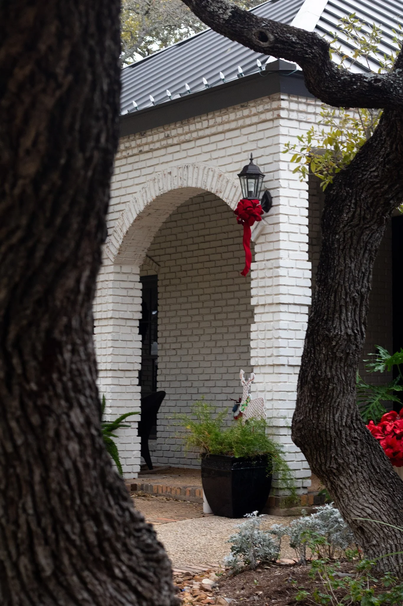A white brick house with an arched porch, decorated for the holidays with a red bow on a black lantern, and holiday figures including a reindeer in a pot. Two large tree trunks frame the scene.