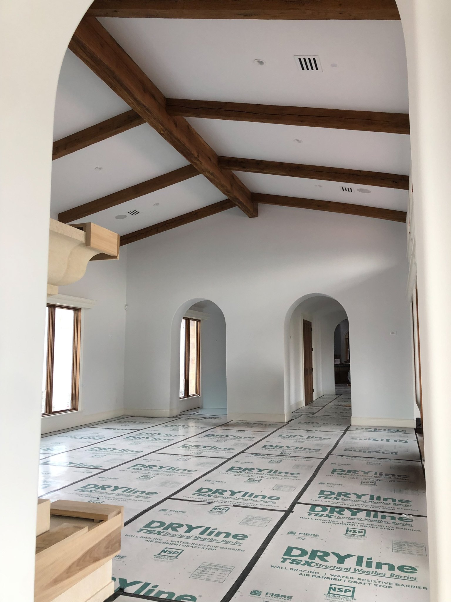 Empty room with white walls, wooden ceiling beams, two windows, and a doorway, covered with protective sheets labeled 'DRYline' during construction.