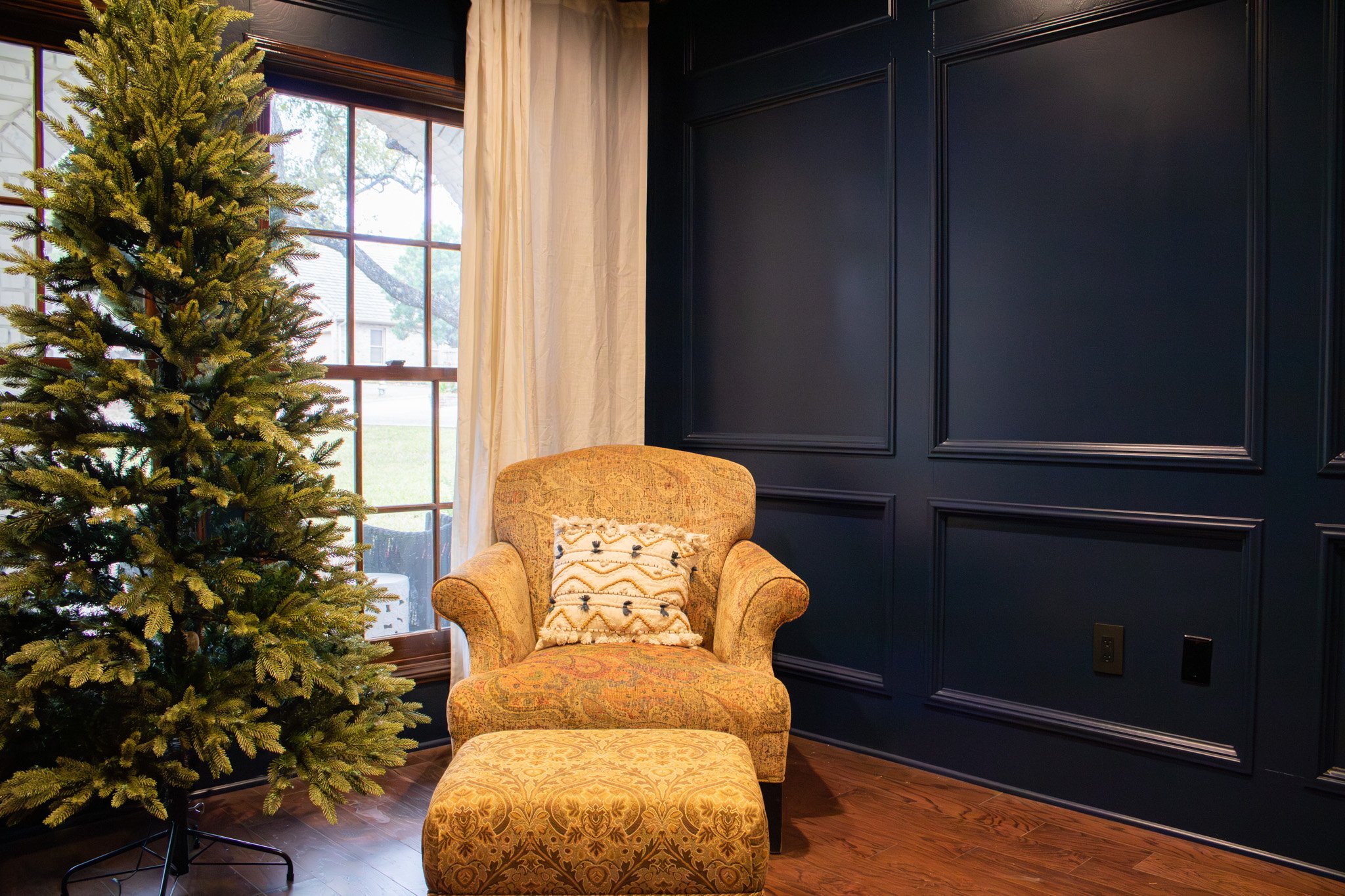A room with a decorated Christmas tree, a large window with white curtains, a beige upholstered armchair with a decorative pillow, and dark blue paneled walls.