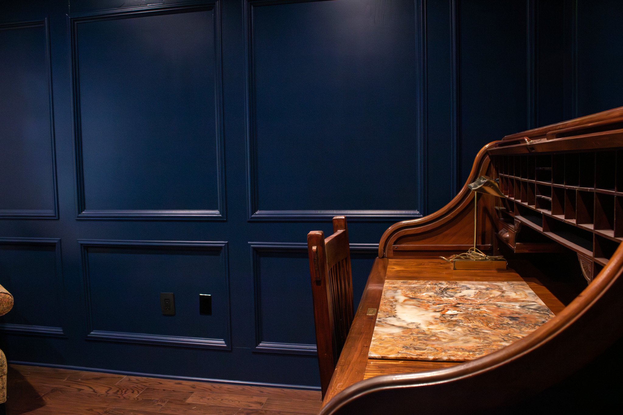 A vintage wooden writing desk with a marble top and an ornate roll-top cover, set against a dark blue paneled wall with a wall outlet and a light switch, in a cozy room.