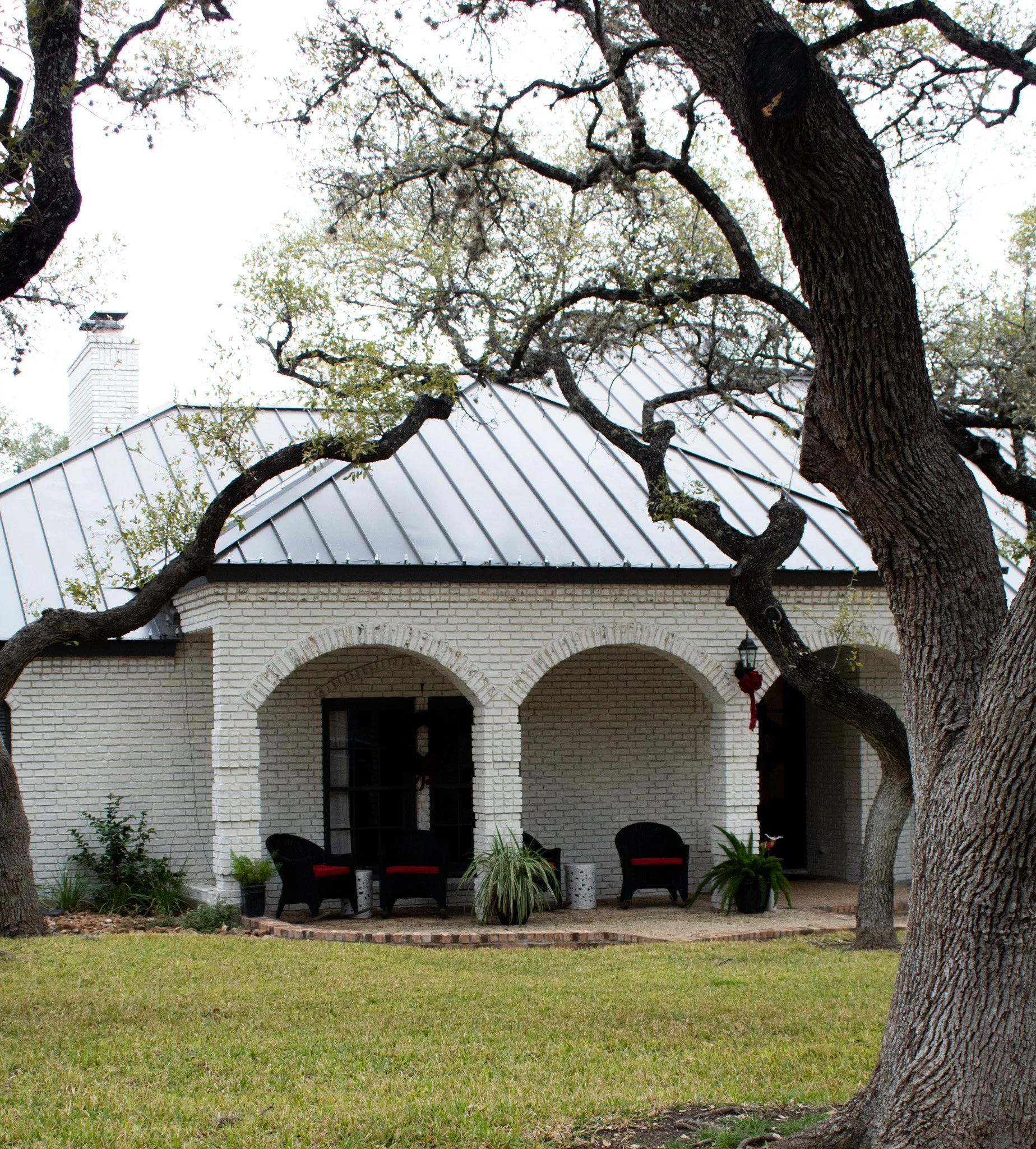 A white brick house with a metal roof, a covered porch, and black chairs with red cushions outside. Large trees with twisting branches surround the house.