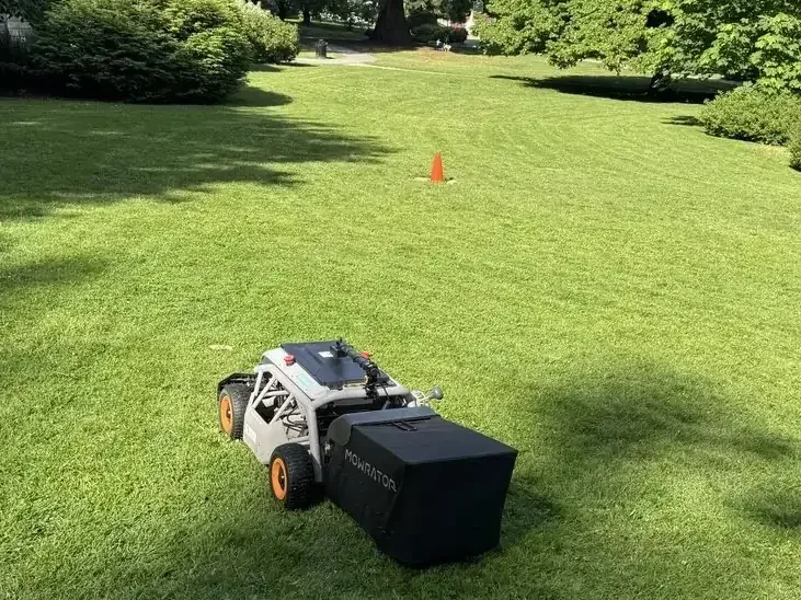 Robotic lawn mower on a green lawn bordered by trees and garden beds.