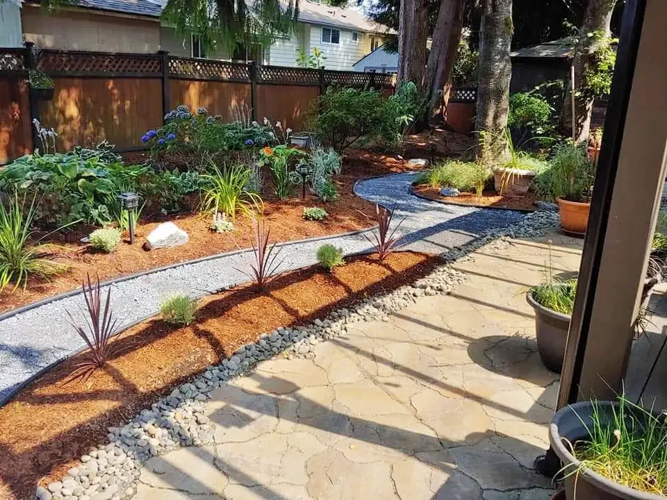 Landscaped garden with stone pathway, potted plants, mulch, and ornamental shrubs against a wooden fence backdrop.