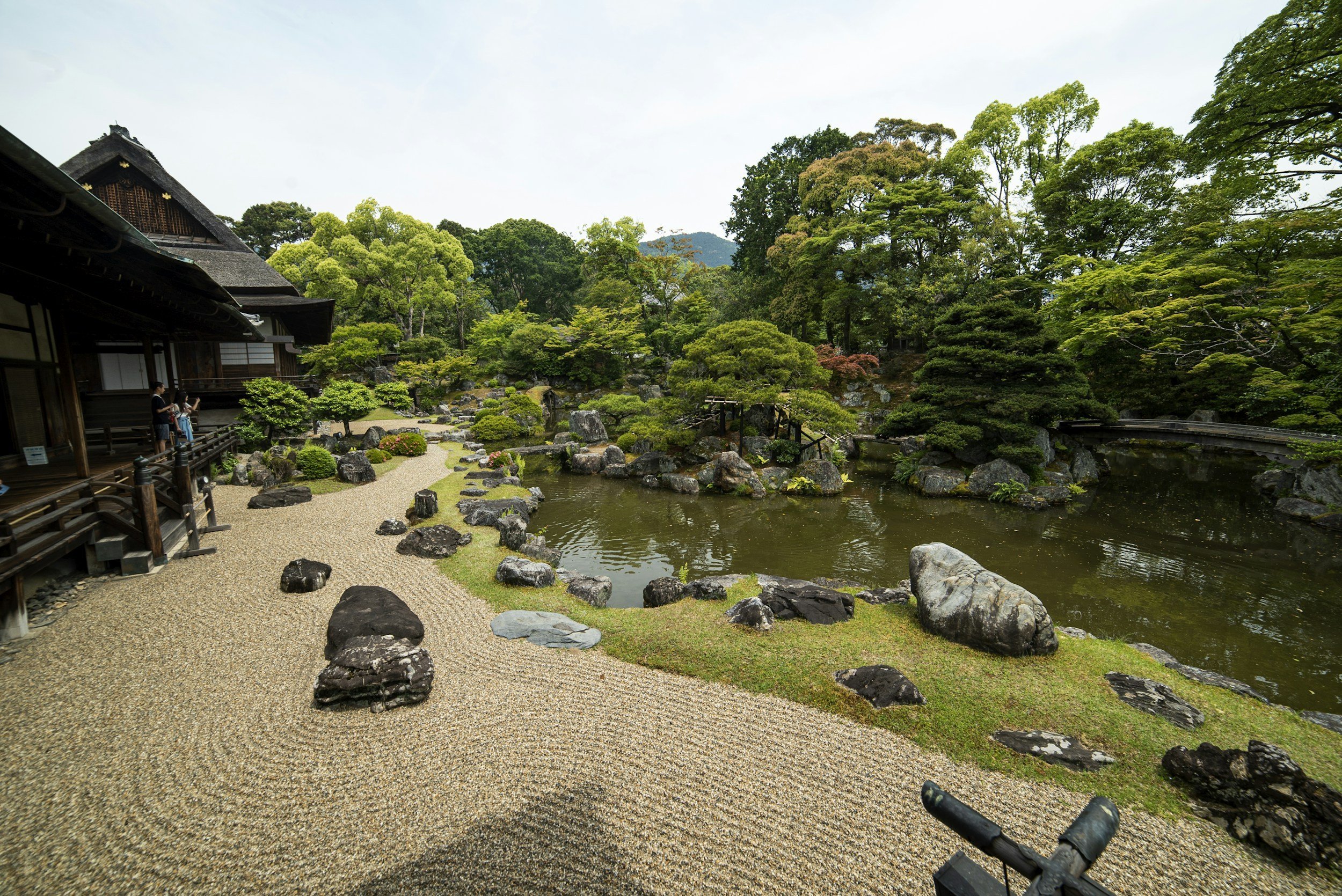 Japanese garden with a pond, raked gravel, moss, and sculpted trees beside a traditional wooden building.