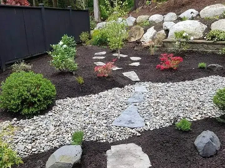 Landscaped garden with stepping stones, mulch, pebbles, shrubs, and trees, bordered by rocks and a black fence, surrounded by forest.