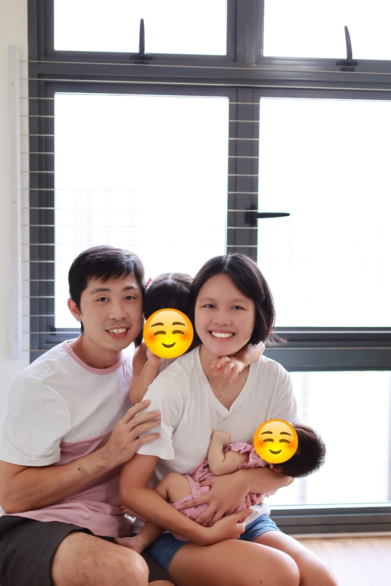 A family of four posing in front of a large window with blinds, smiling and embraces. The children are wearing casual clothes, and their faces are covered with smiley face emojis.