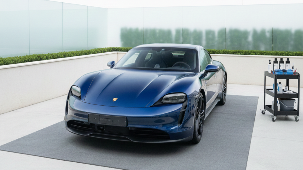 A blue sports car parked on a gray mat on a rooftop or outdoor terrace with a cleaning cart nearby.
