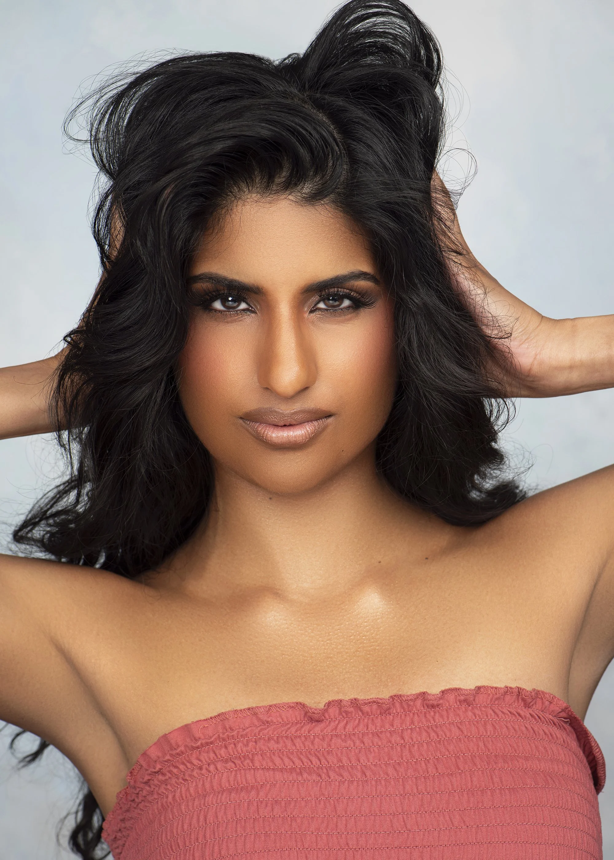 Close-up of a woman with dark wavy hair, makeup, and a strapless reddish-orange top, posing with hands behind her head.