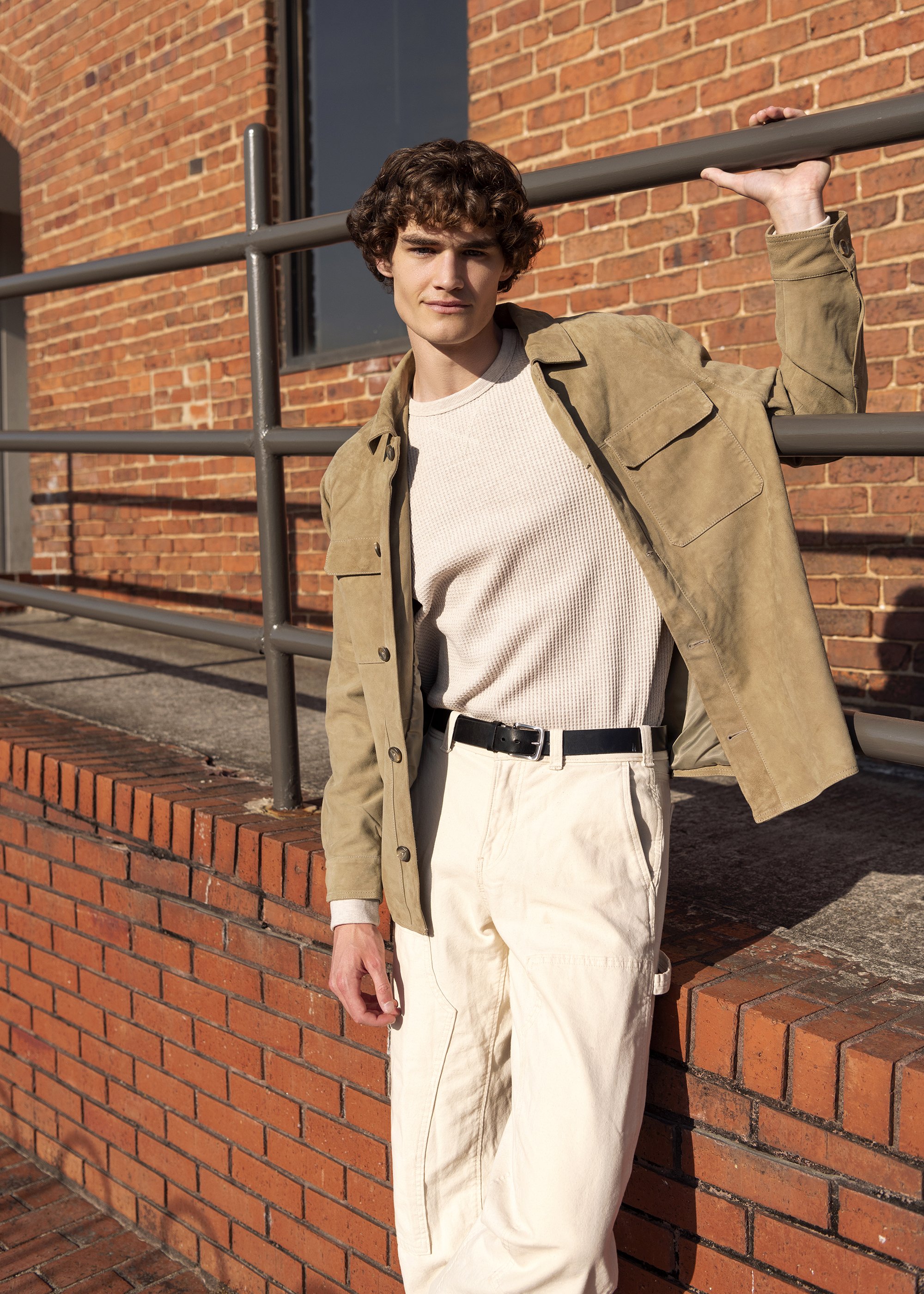 A young man with curly brown hair leaning against a metal railing outdoors in front of a brick building, wearing a beige jacket, cream sweater, and cream pants, with sunlight casting shadows.